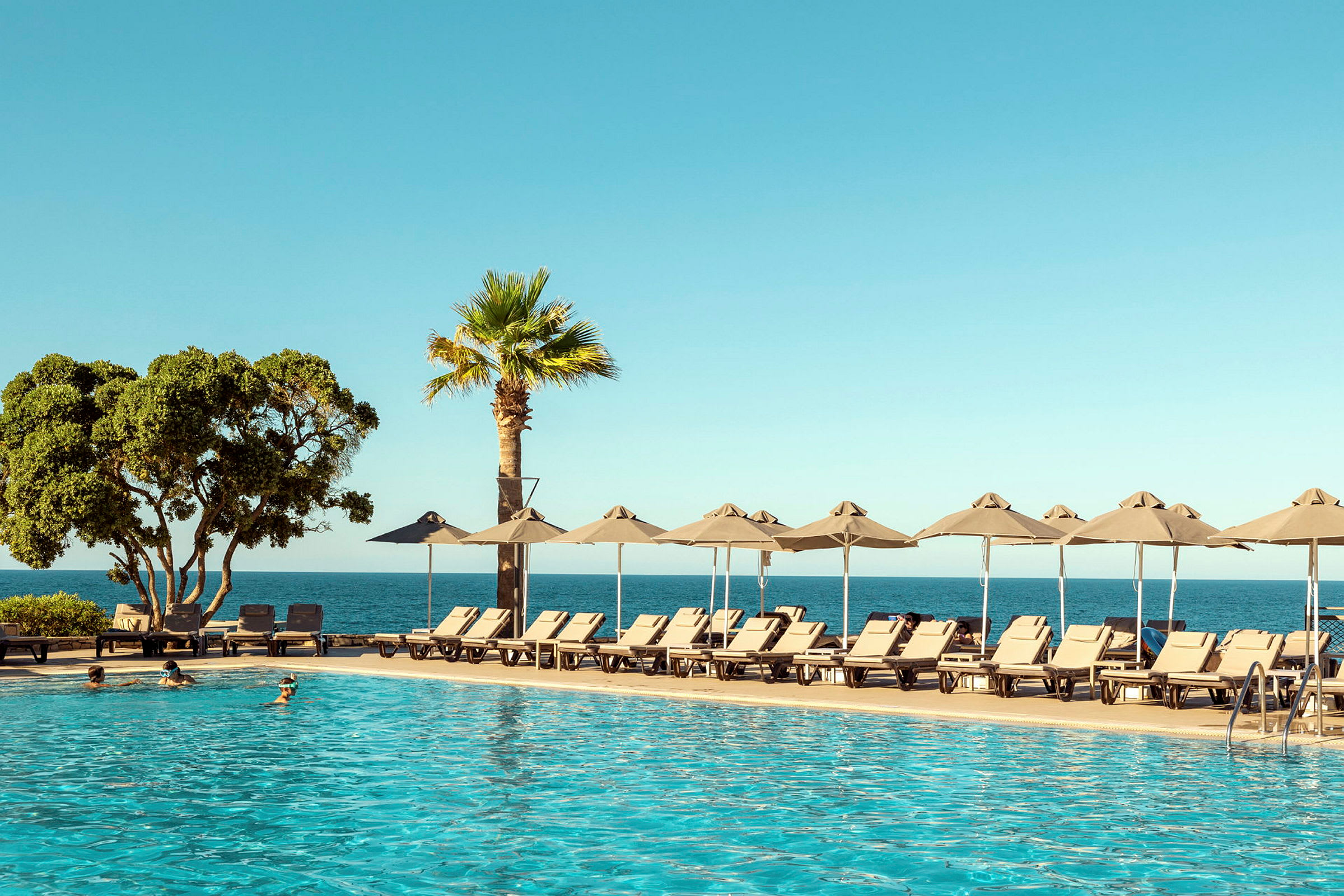 En pool i förgrunden med solstolar och parasoller vid sidan. Bakom syns havet och en klar blå himmel. (AI-genererad text)