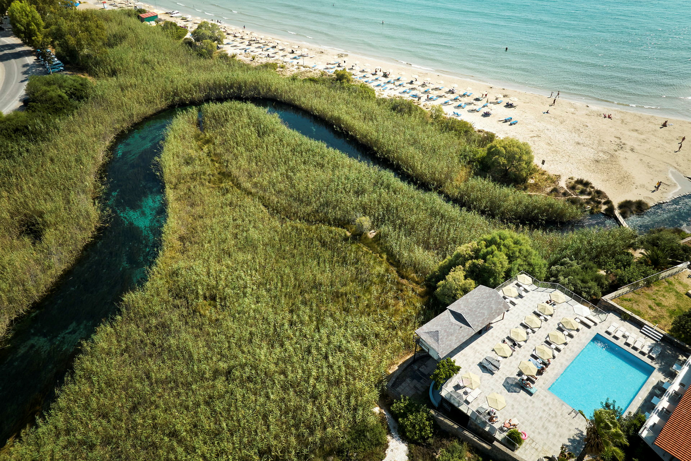 Flygvy av ett landskap med en pool, en strand med solstolar och ett vattendrag omgivet av vegetation. (AI-genererad text)