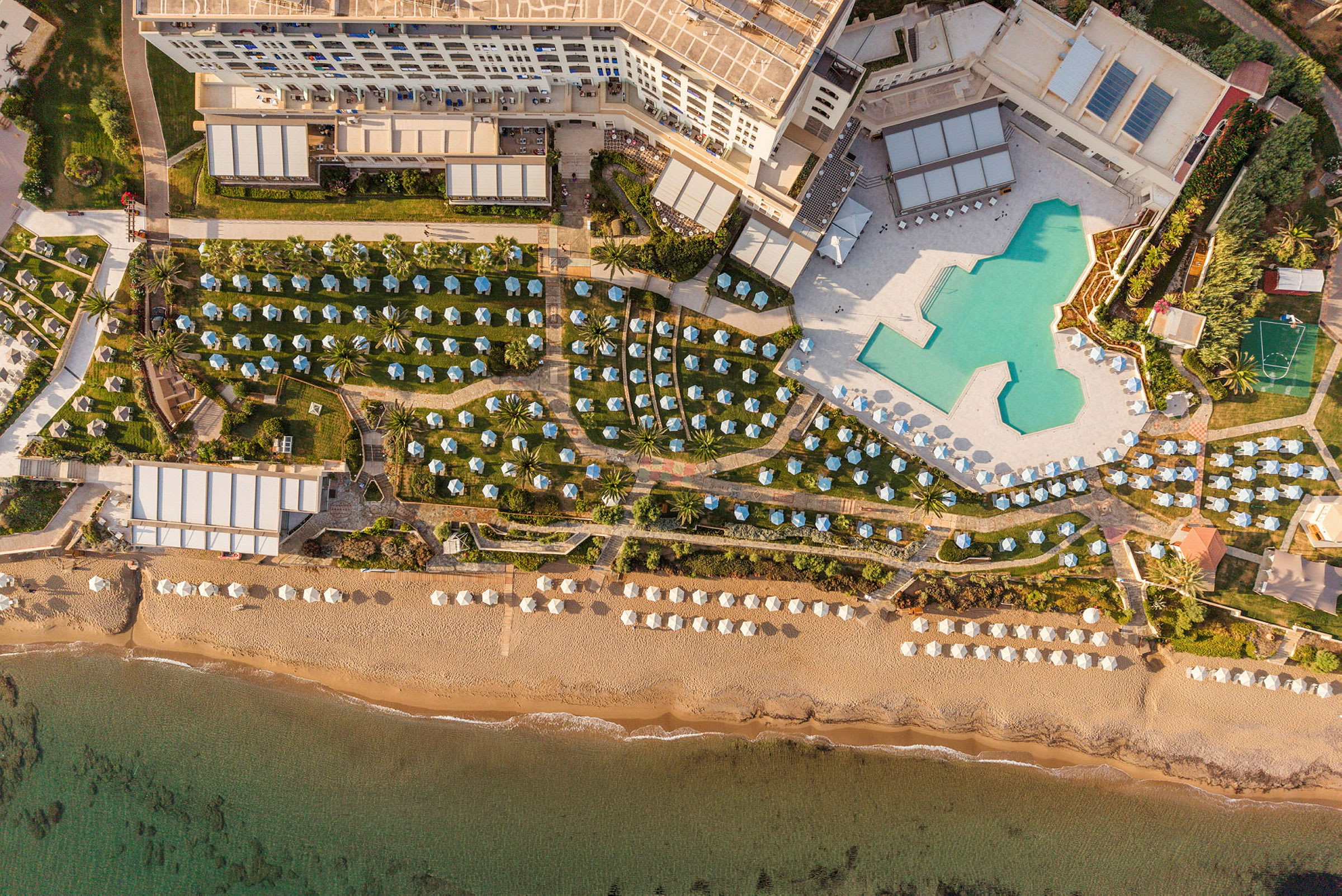 Flygbild av en resort med stora poolområden, gröna gräsmattor med solstolar och parasoller, samt en sandstrand vid havet. (AI-genererad text)