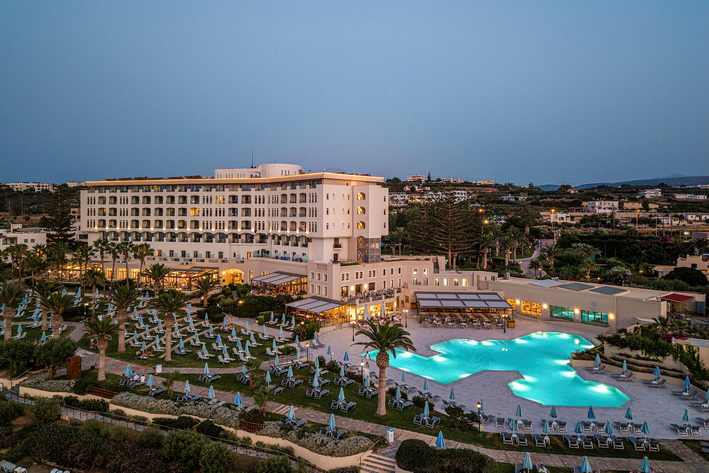 Hotellbyggnad med flera våningar, omgiven av en upplyst pool och solstolar, omgärdat av grönska och omgivande byggnader vid skymning. (AI-genererad text)