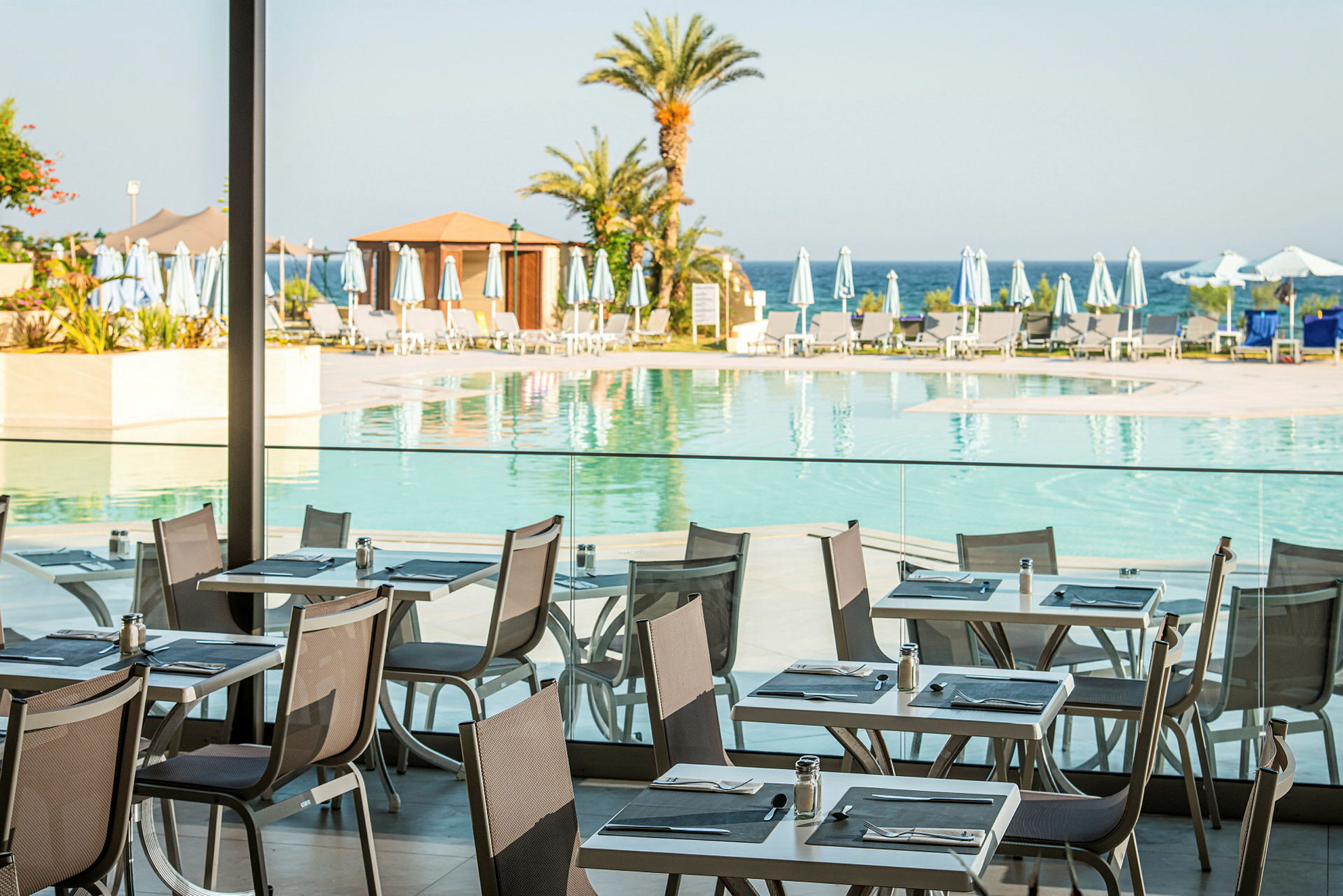 Motiv av ett utomhusområde med en pool vid en restaurang med bord och stolar, omgivna av solstolar och parasoller. Palmer syns och havet i bakgrunden. (AI-genererad text)