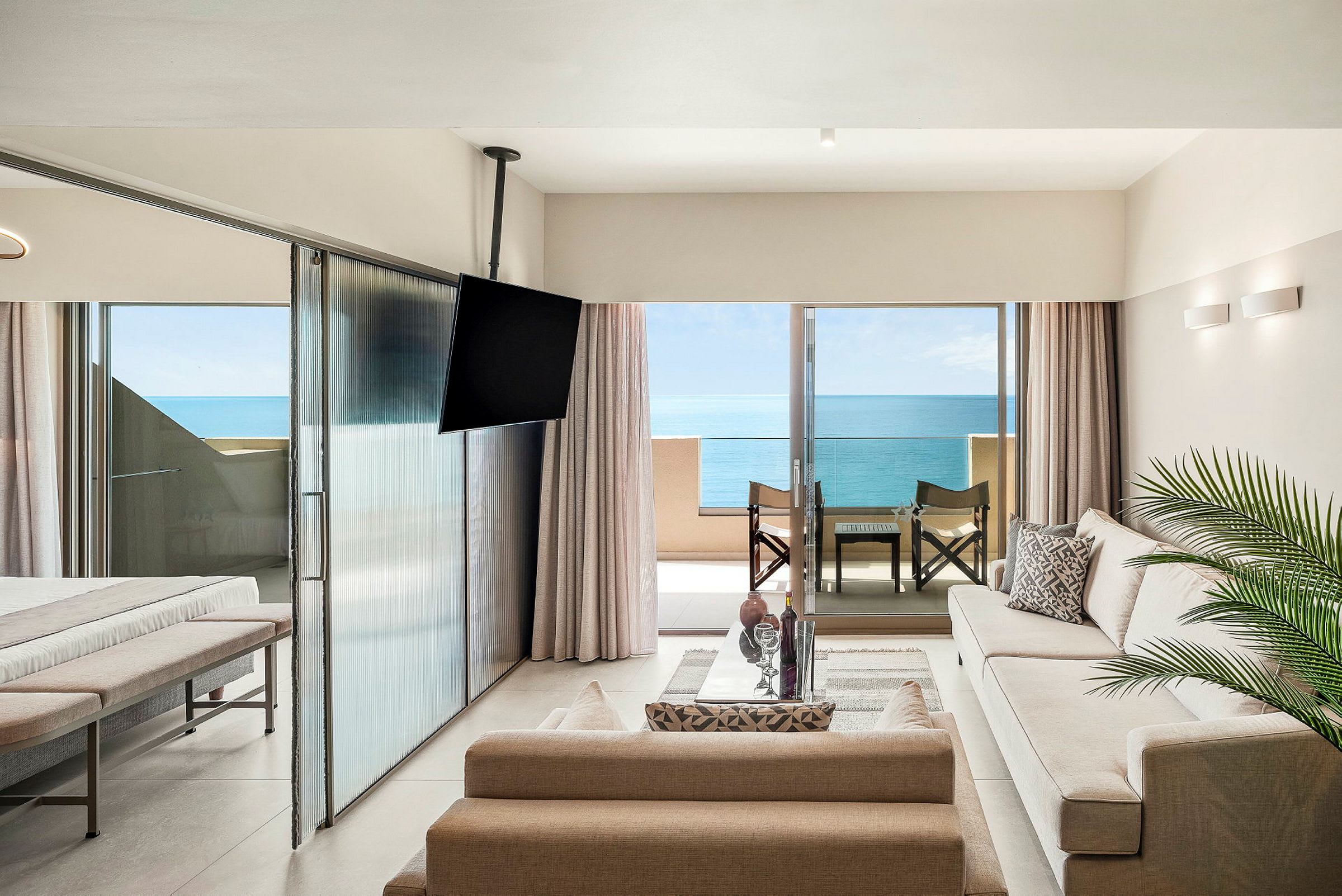 Ett modernt hotellrum med sittgrupp, TV och havsutsikt från en balkong med sittplats. (AI-genererad text)