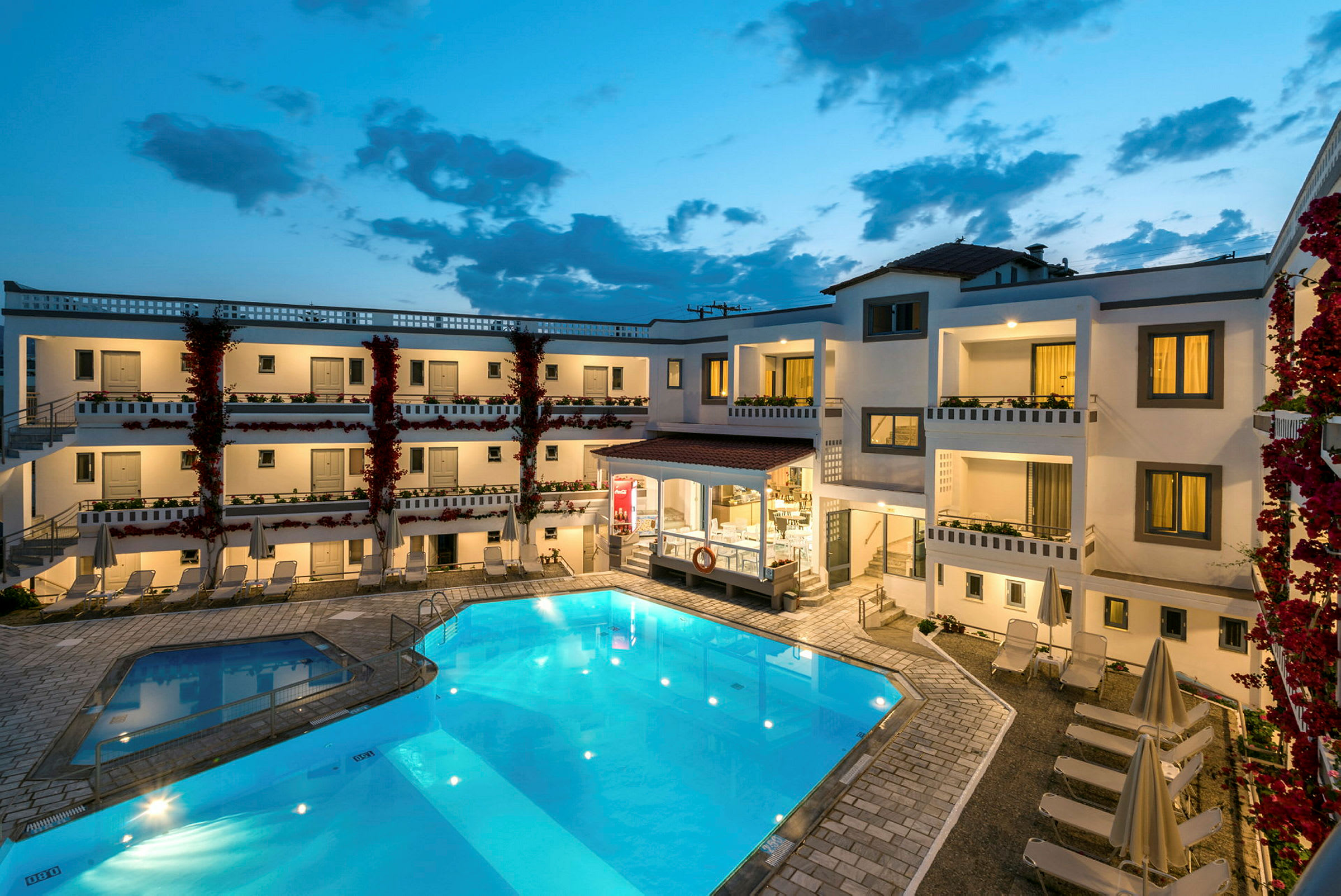 Bild av ett hotellområde med en upplyst pool, solstolar och omgivande byggnader med balkonger under kvällstid. (AI-genererad text)