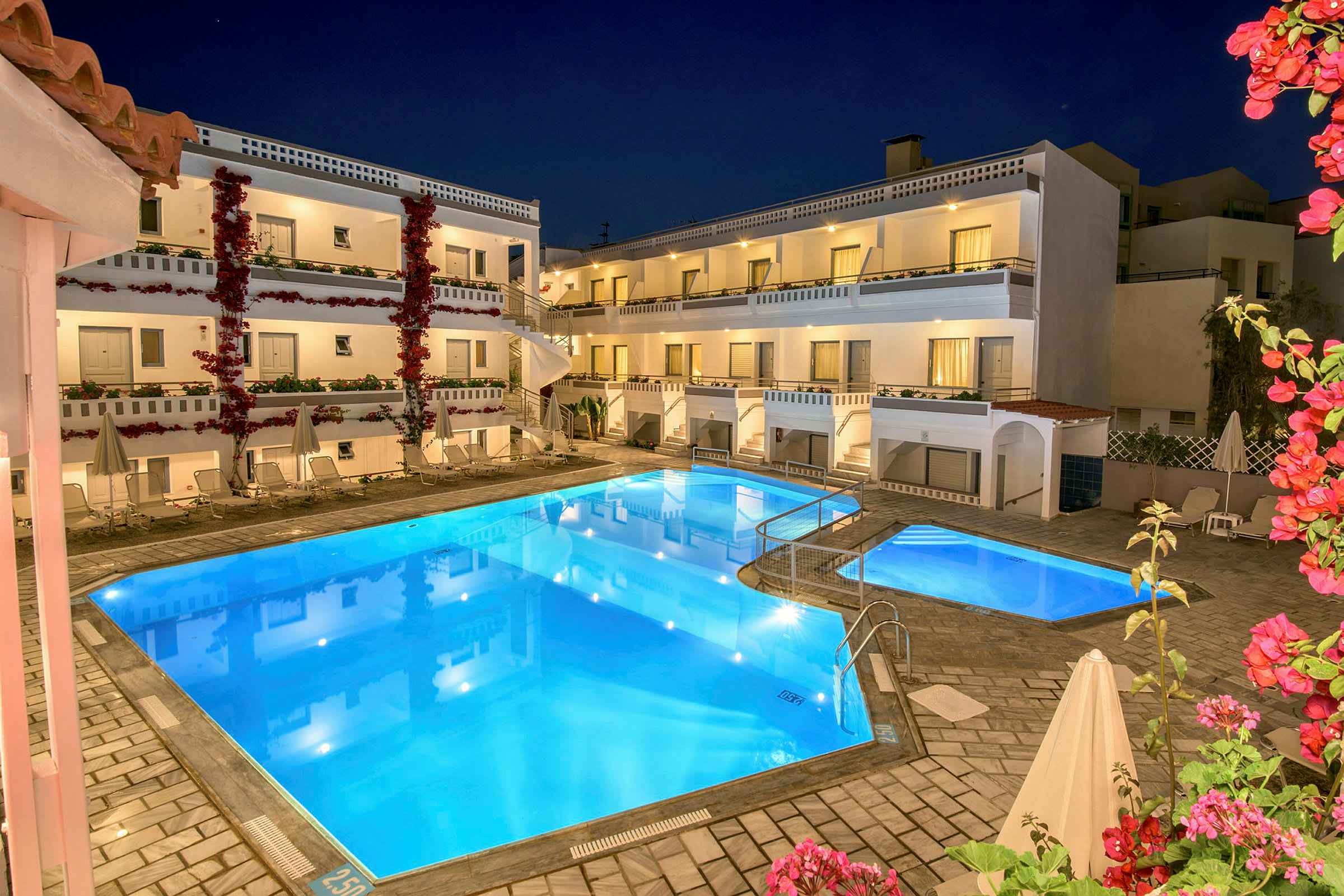 Hotell med tre våningar, pool i förgrunden och blommande växter. (AI-genererad text)