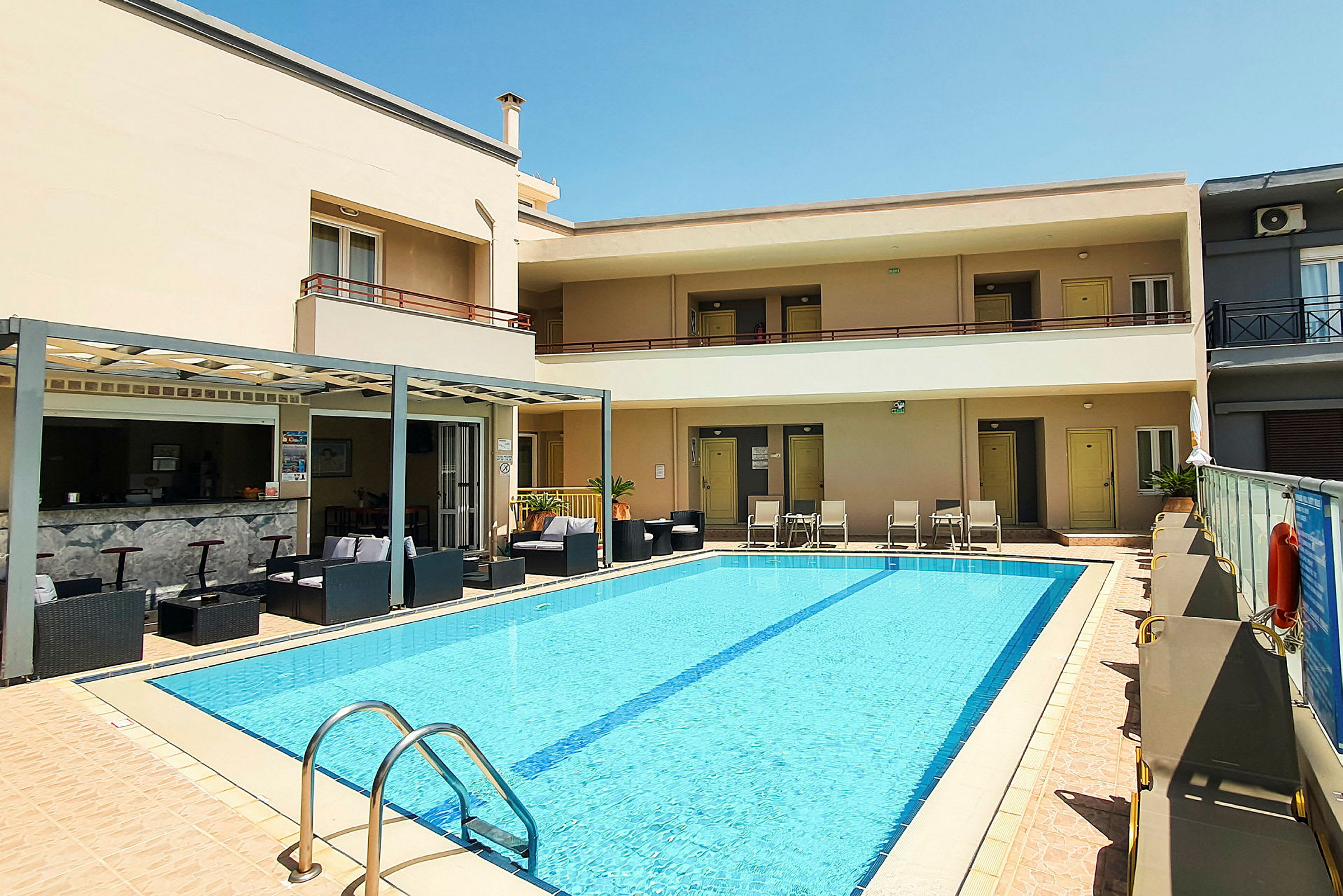 Bild av ett hotell med en pool, omgiven av solstolar. En bar och sittplatser syns i bakgrunden. (AI-genererad text)