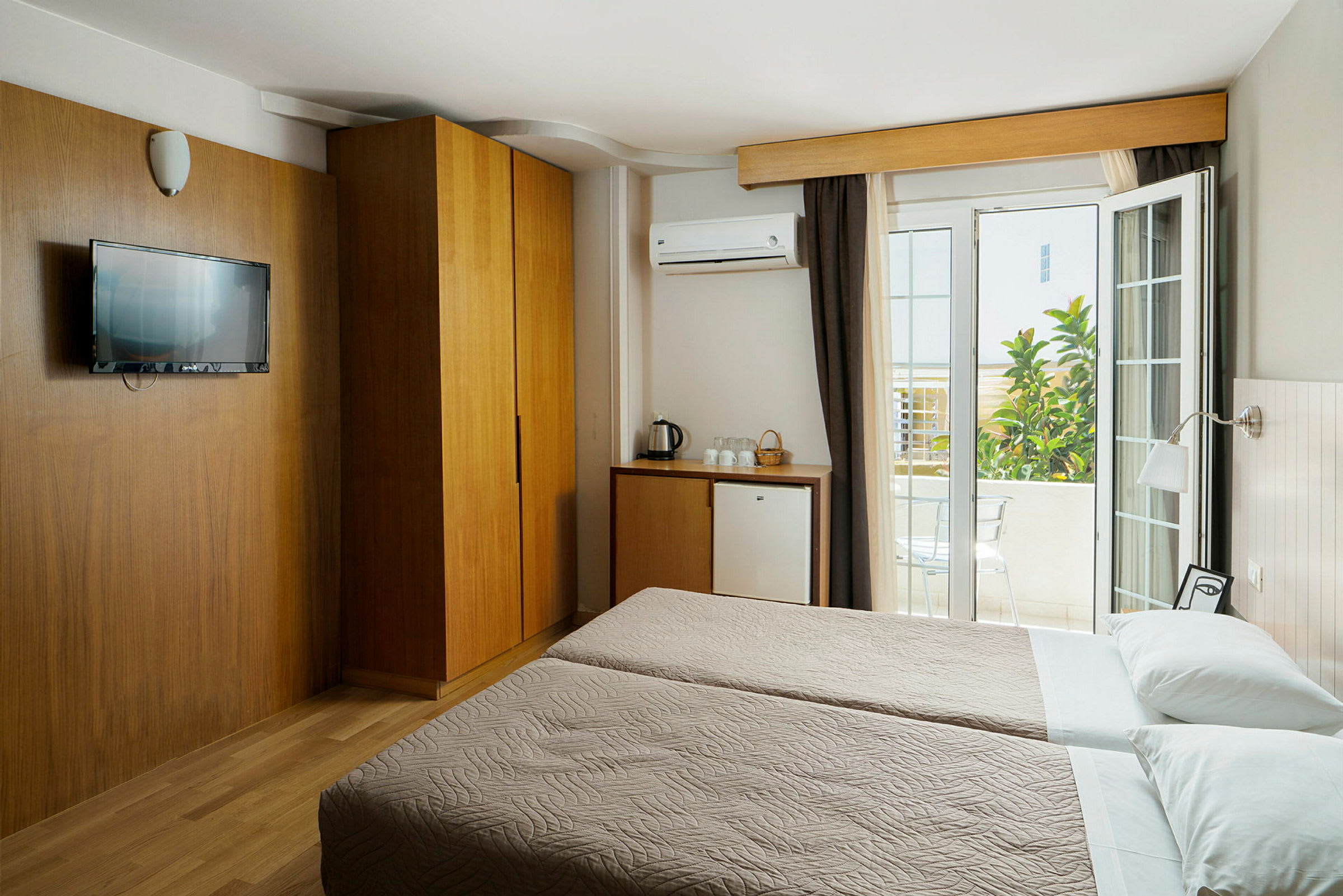 Hotellrum med två enkelsängar, en träskåp, kylskåp och en öppen balkongdörr. (AI-genererad text)