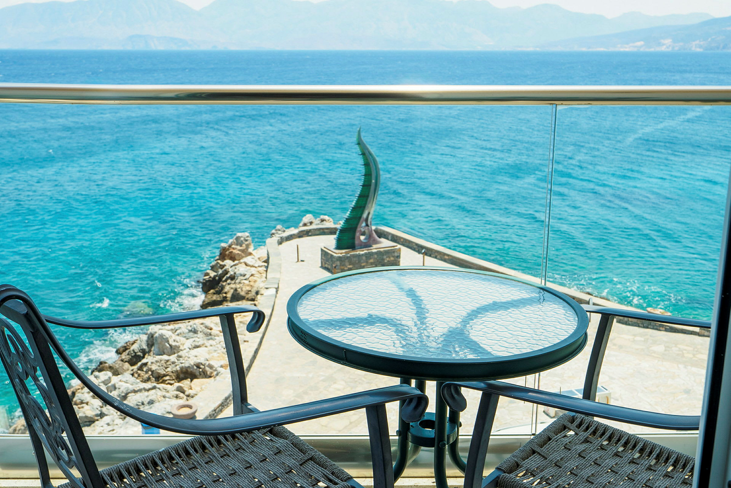 Utsikt från en terrass mot havet med två stolar och ett bord i förgrunden. (AI-genererad text)