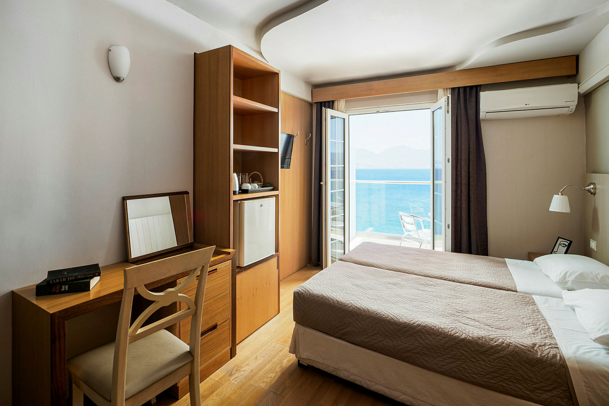 Ett hotellrum med två enkelsängar, en skrivbord och stol, samt ett skåp med kylskåp. Utanför syns en balkong med havsutsikt. (AI-genererad text)