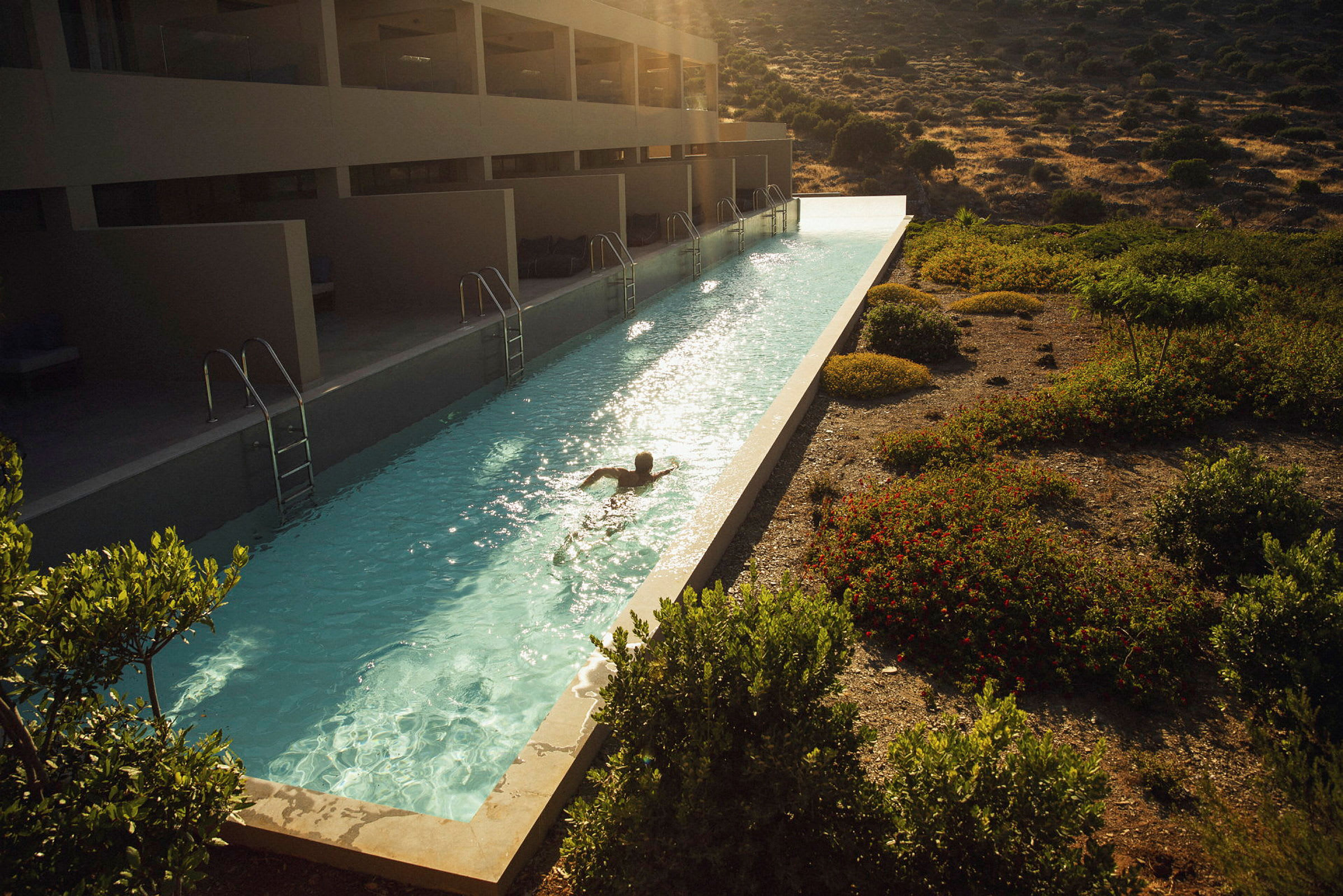 Person simmar i långsmal pool vid hotellbyggnad, omgivande grönska och en bergigt landskap. (AI-genererad text)