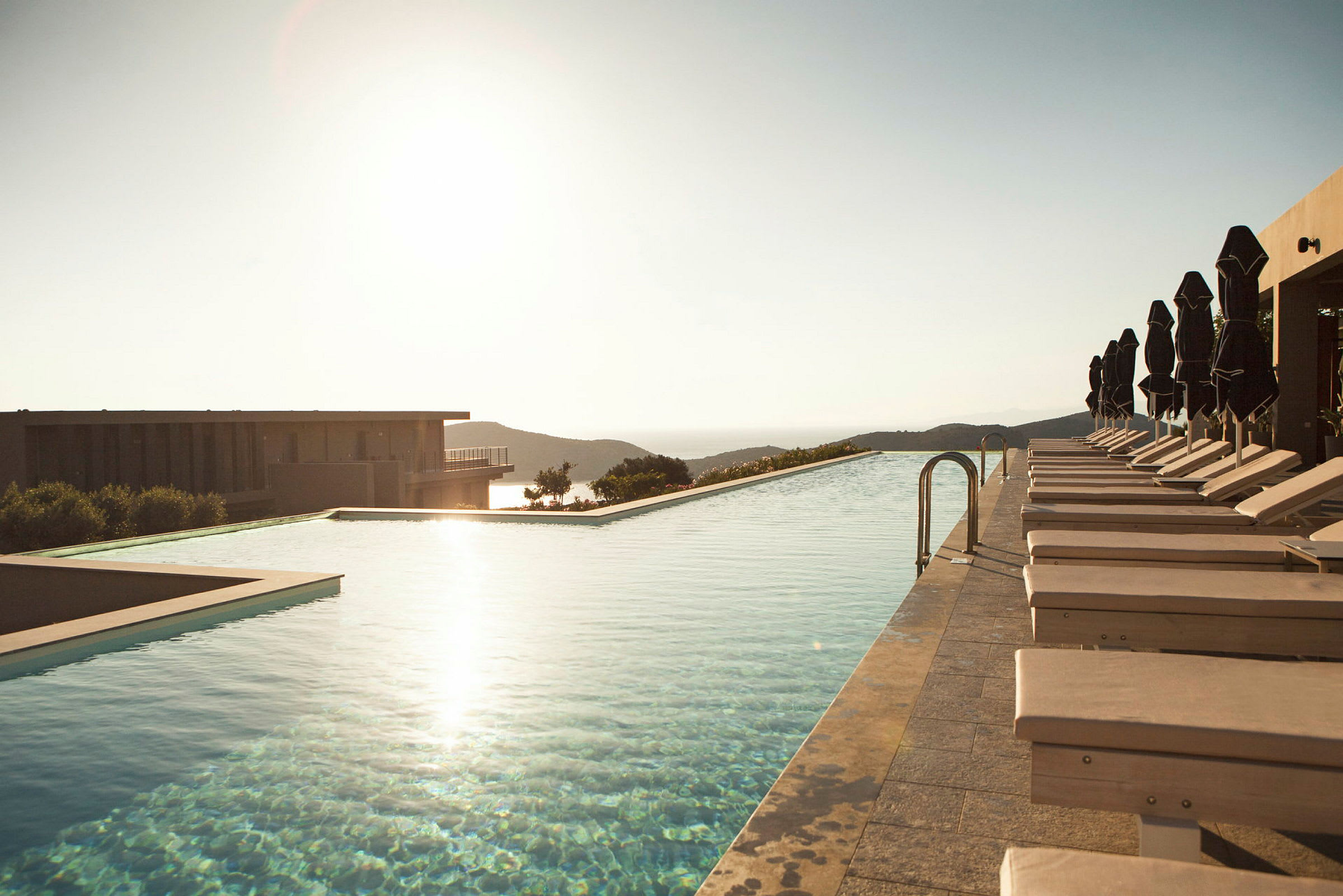 En infinitypool med solstolar och parasoller bredvid, med utsikt över kullar och hav vid solnedgång. (AI-genererad text)