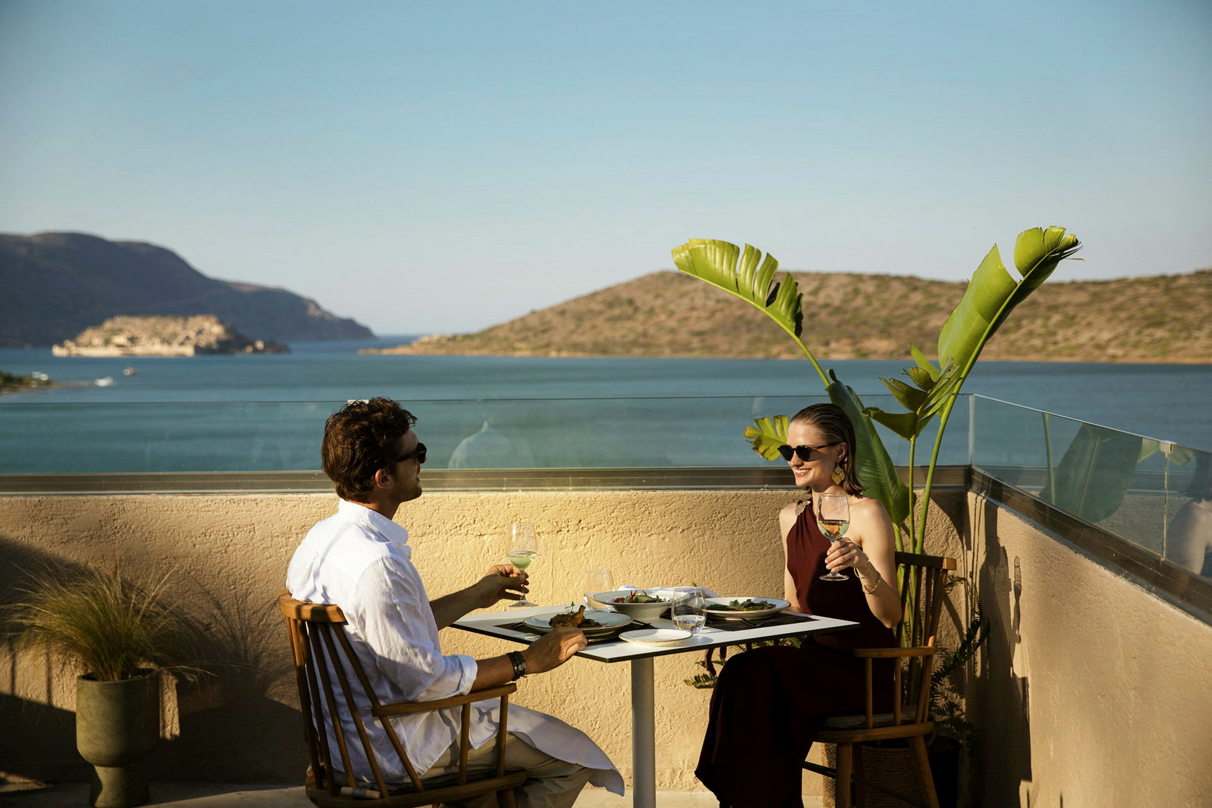 Två personer sitter vid ett bord med mat och dryck på en terrass med utsikt över havet och berg. (AI-genererad text)