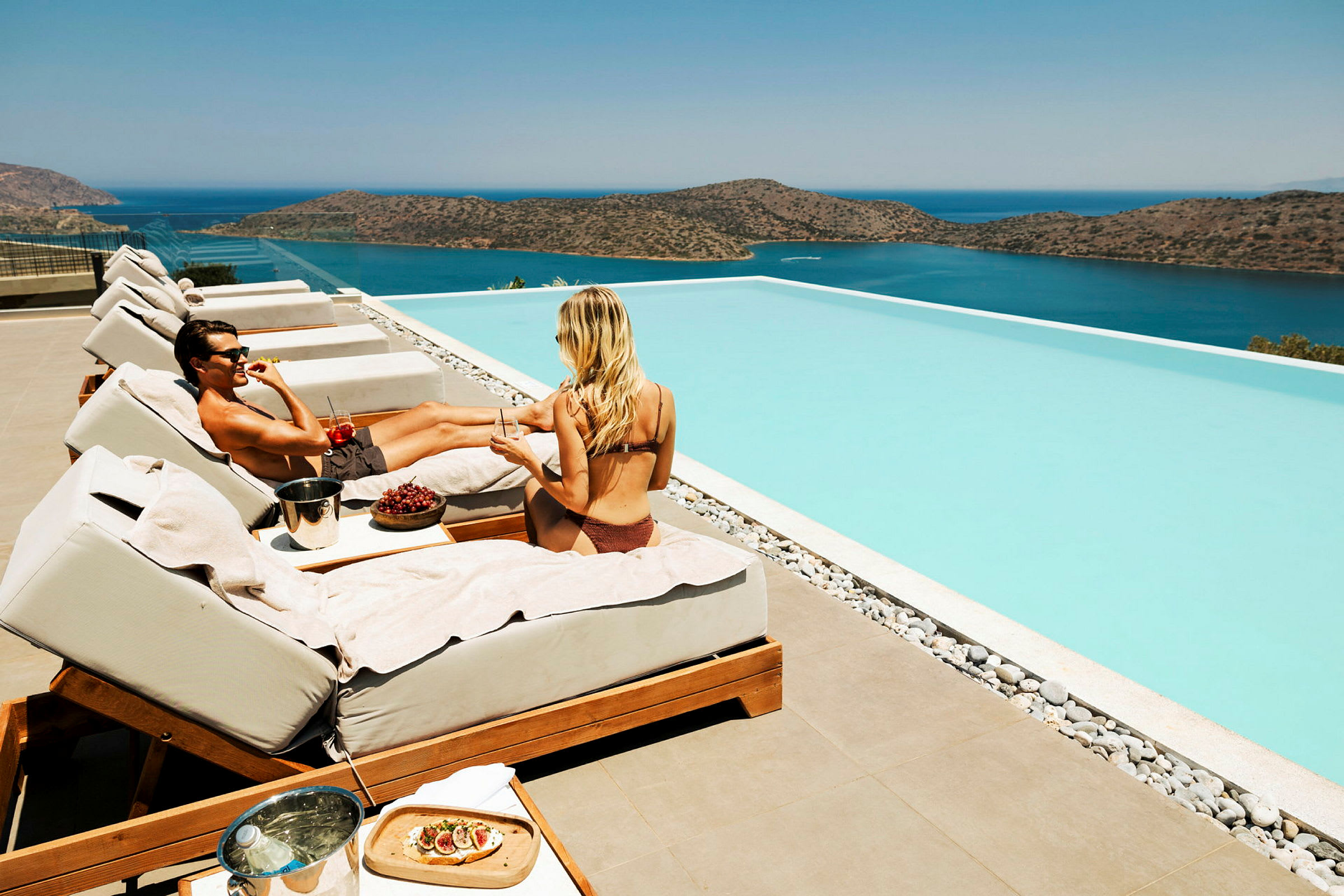 Två personer sitter på solstolar vid en infinitypool och en sjö utsikt. (AI-genererad text)