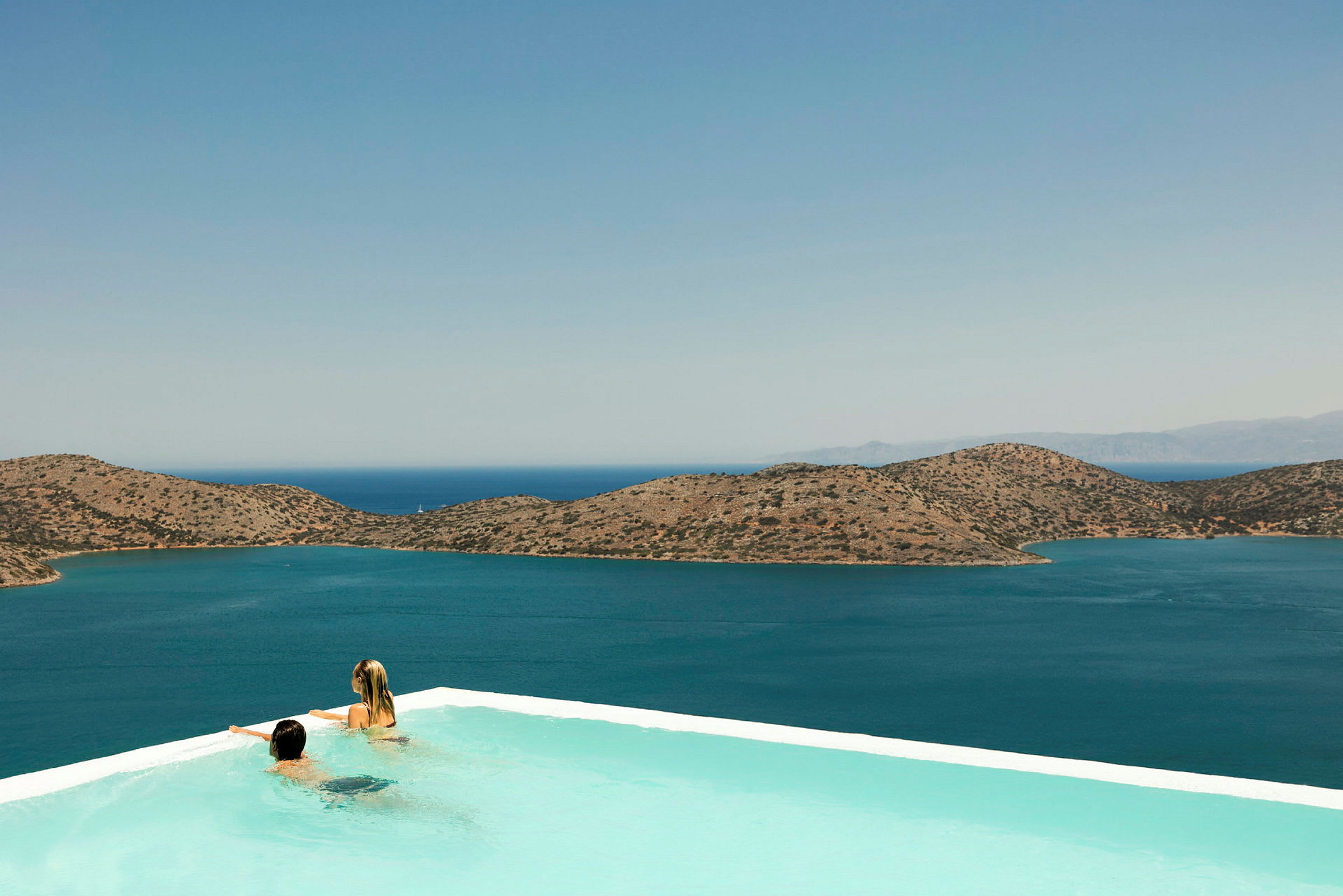 Två personer i en infinitypool med utsikt över havet och berg i bakgrunden. (AI-genererad text)