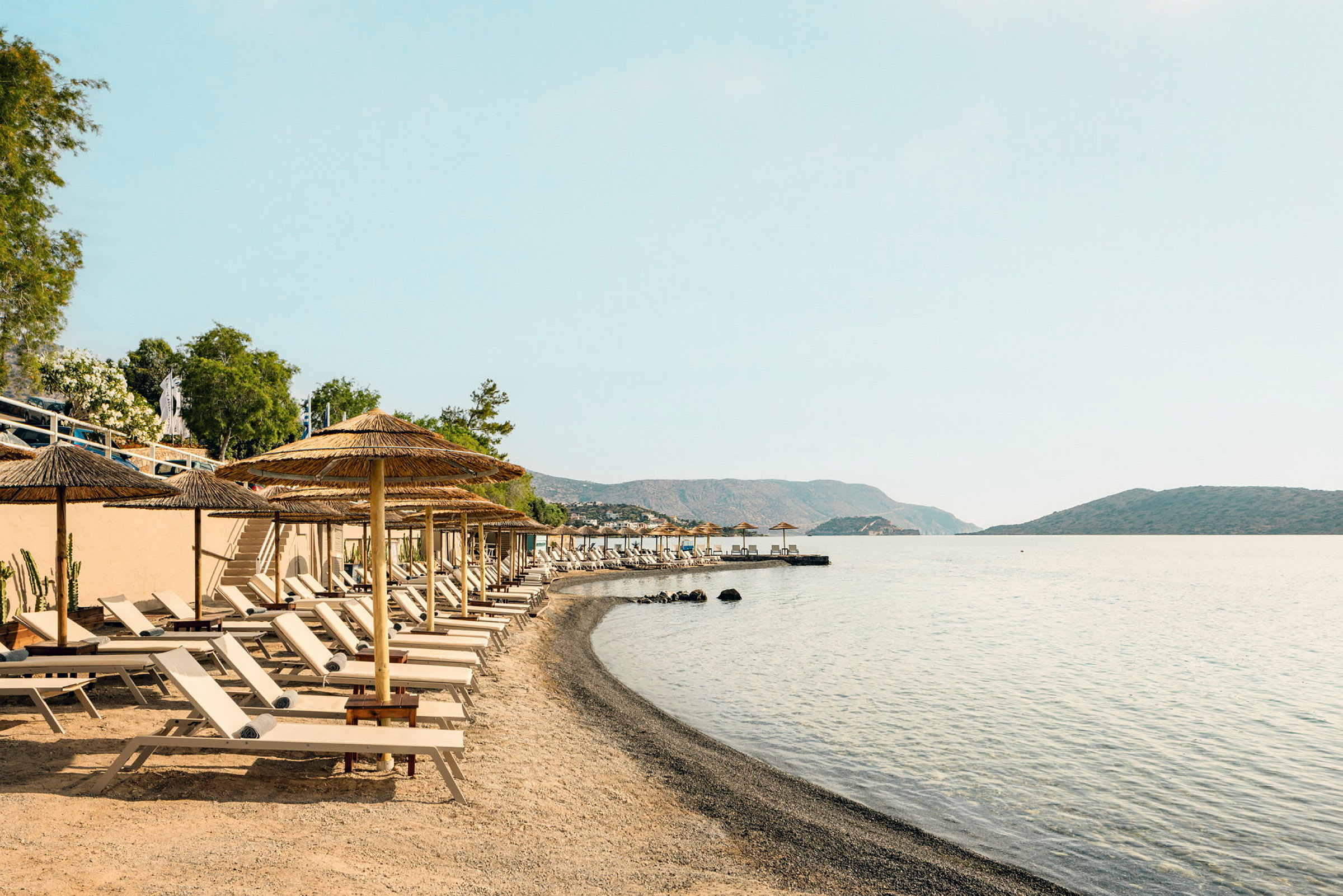 Strand med solstolar och parasoller vid havet, omgiven av berg och träd. (AI-genererad text)
