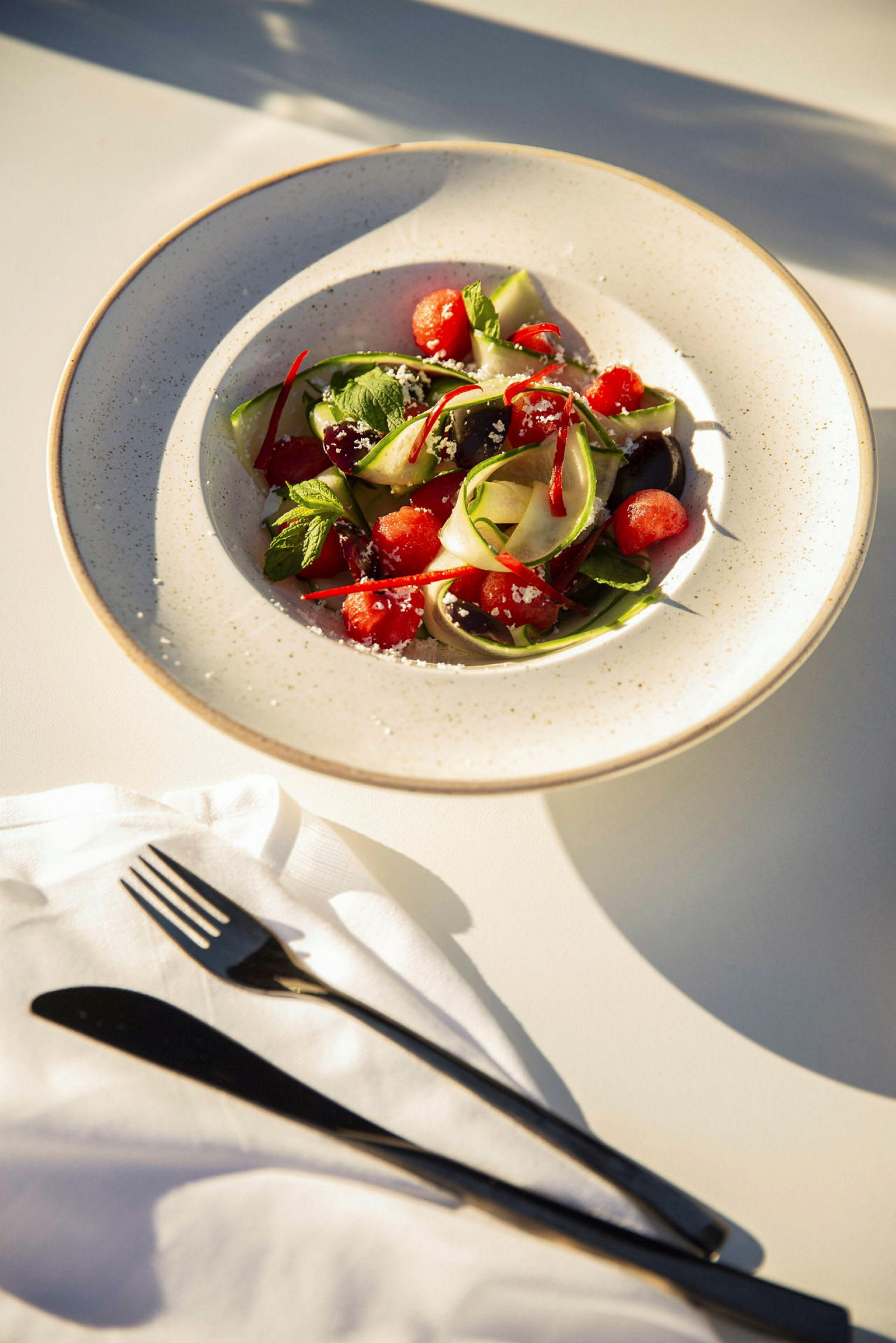 En skål med vattenmelon, zucchini och örter på ett bord med bestick. (AI-genererad text)