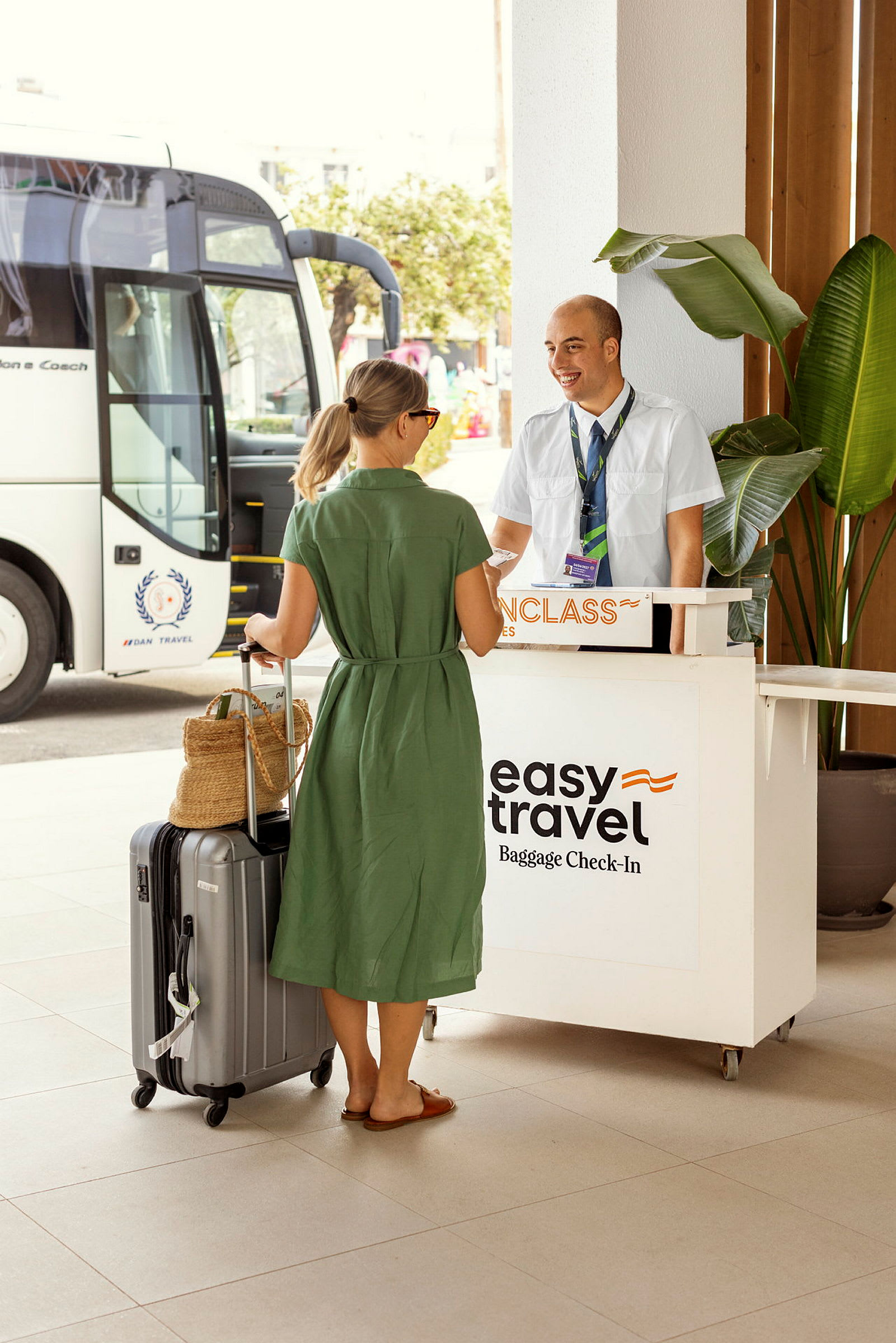 En kvinna står vid en receptionsdisk med en resväska och en bag, nära en resebuss. Skylten på disken indikerar bagageincheckning. (AI-genererad text)