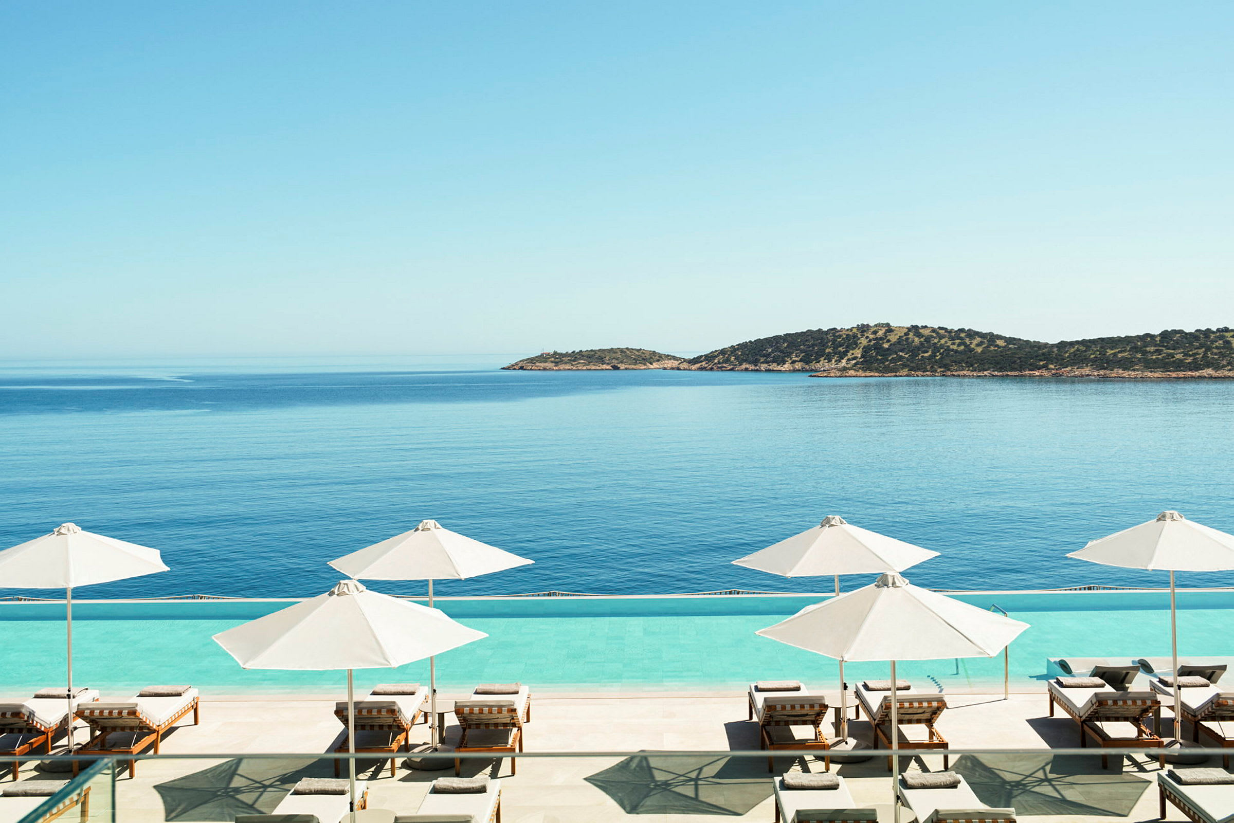 Solstolar och parasoller vid en pool med utsikt över havet och en ö i bakgrunden. (AI-genererad text)