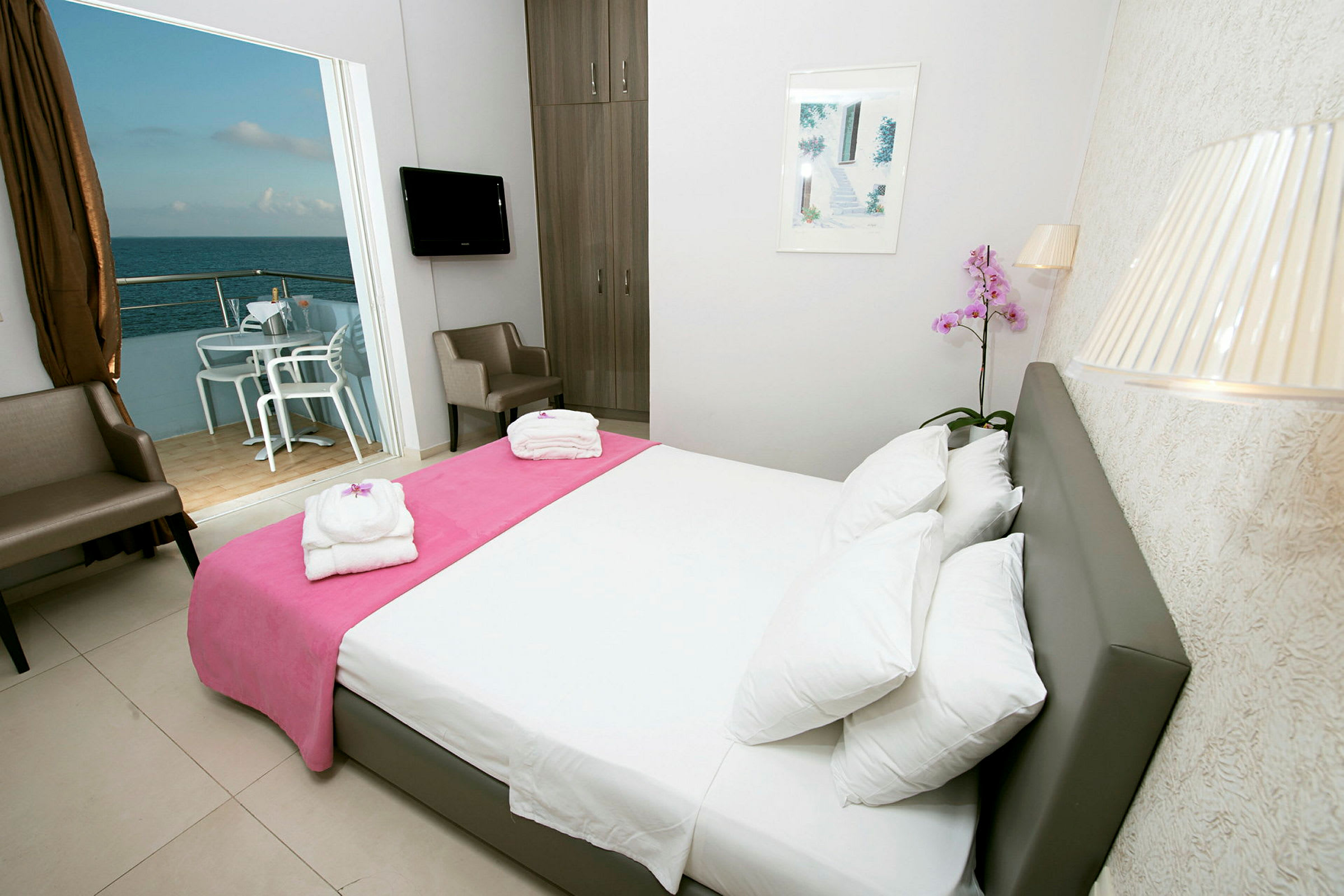 Ett hotellrum med vita väggar, en dubbel säng med en rosa kudde, två stolar, en TV och en balkong med utsikt över havet. (AI-genererad text)