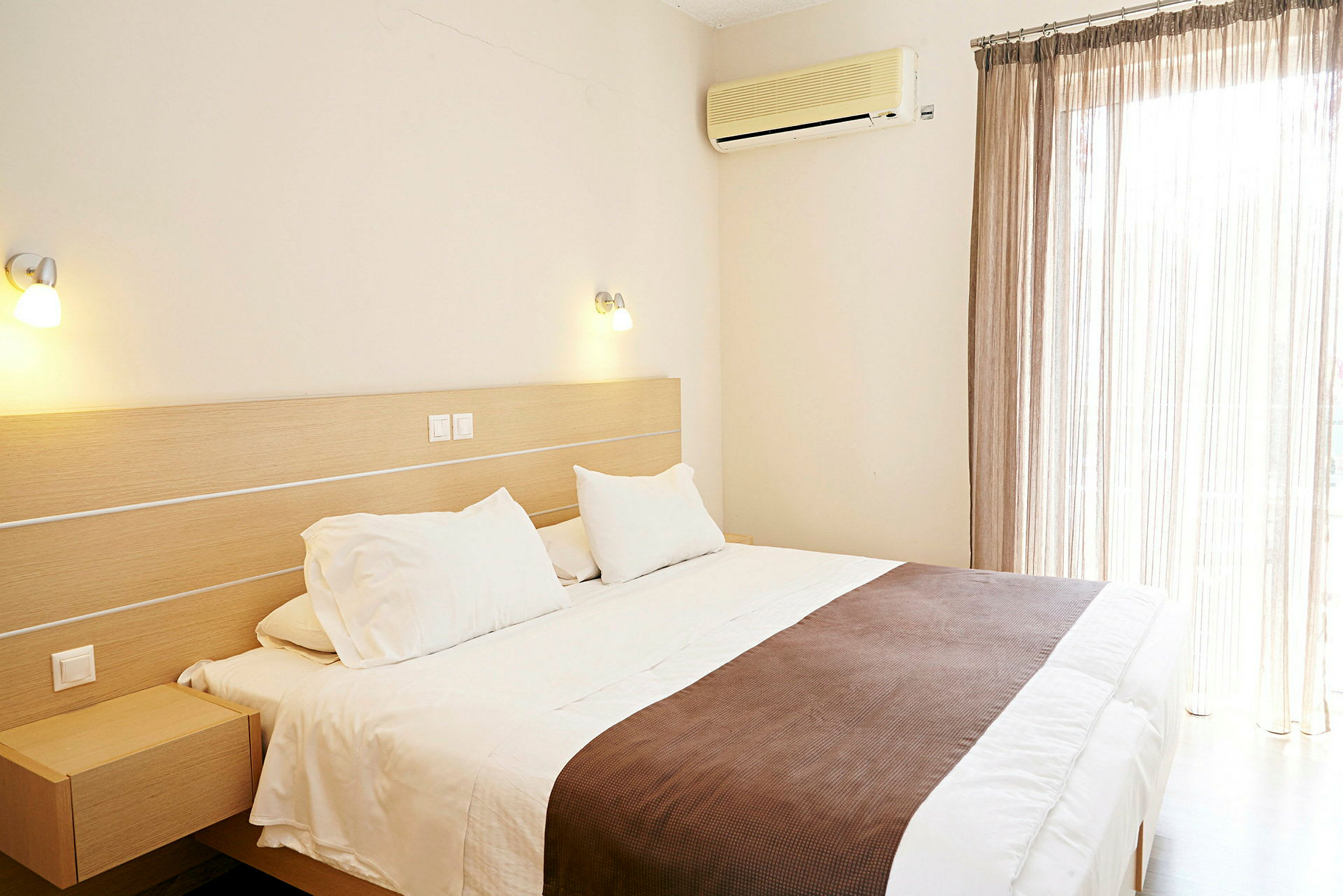 Ett hotellrum med en stor säng, vita sängkläder och brunt överkast. En luftkonditionering är monterad på väggen bredvid fönstret med ljusa gardiner. (AI-genererad text)