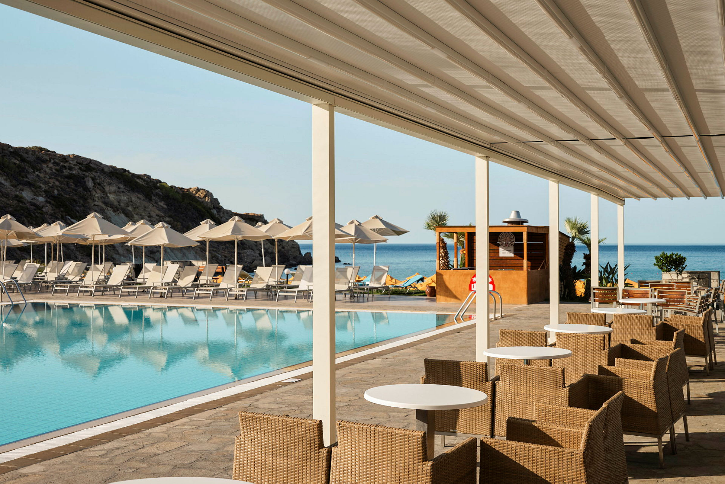 Bild av en poolområde vid en resort, med solstolar och parasoller framför havet. Före poolen finns sittplatser med bord och stolar, täckt av en överbyggnad. (AI-genererad text)