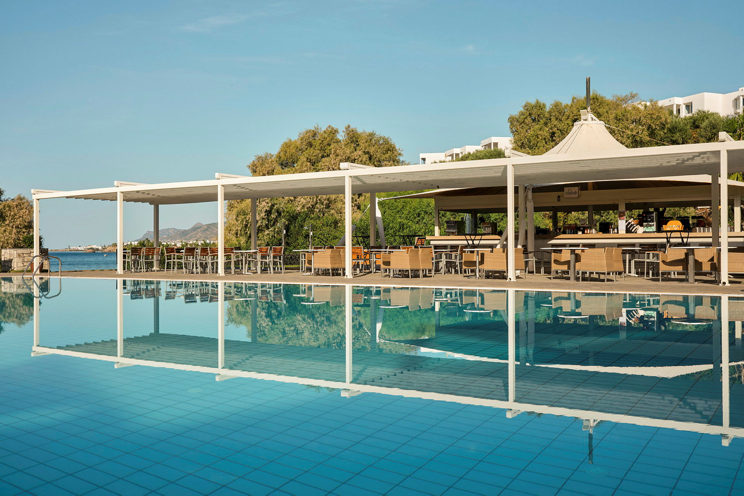 Bild av en pool med en tillhörande sittplats och bar under en vit pergola. I bakgrunden syns grönskande träd och hav. (AI-genererad text)