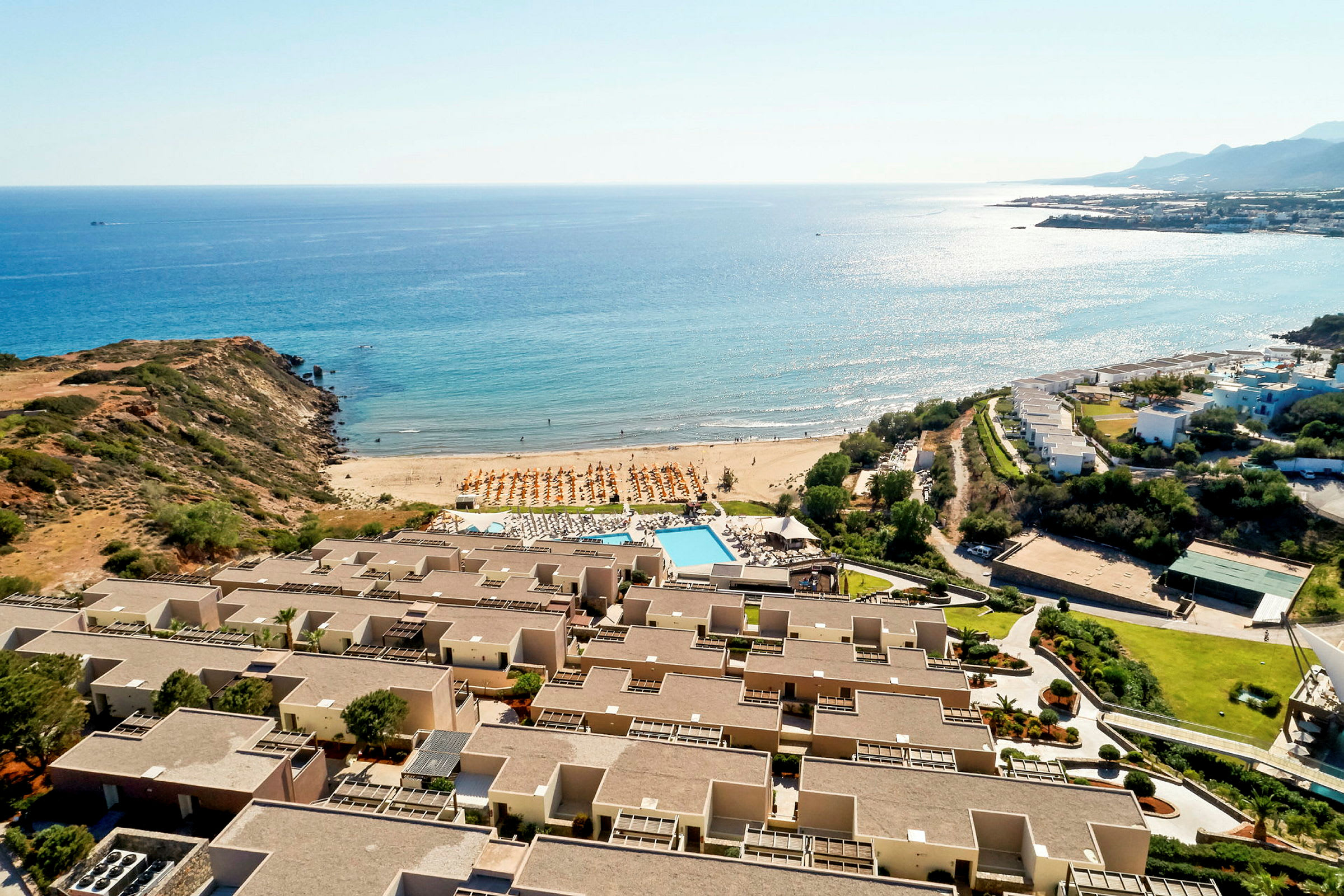 Flygfoto över hotellkomplex nära havet med en sandstrand och parasoller, samt poolområde. (AI-genererad text)