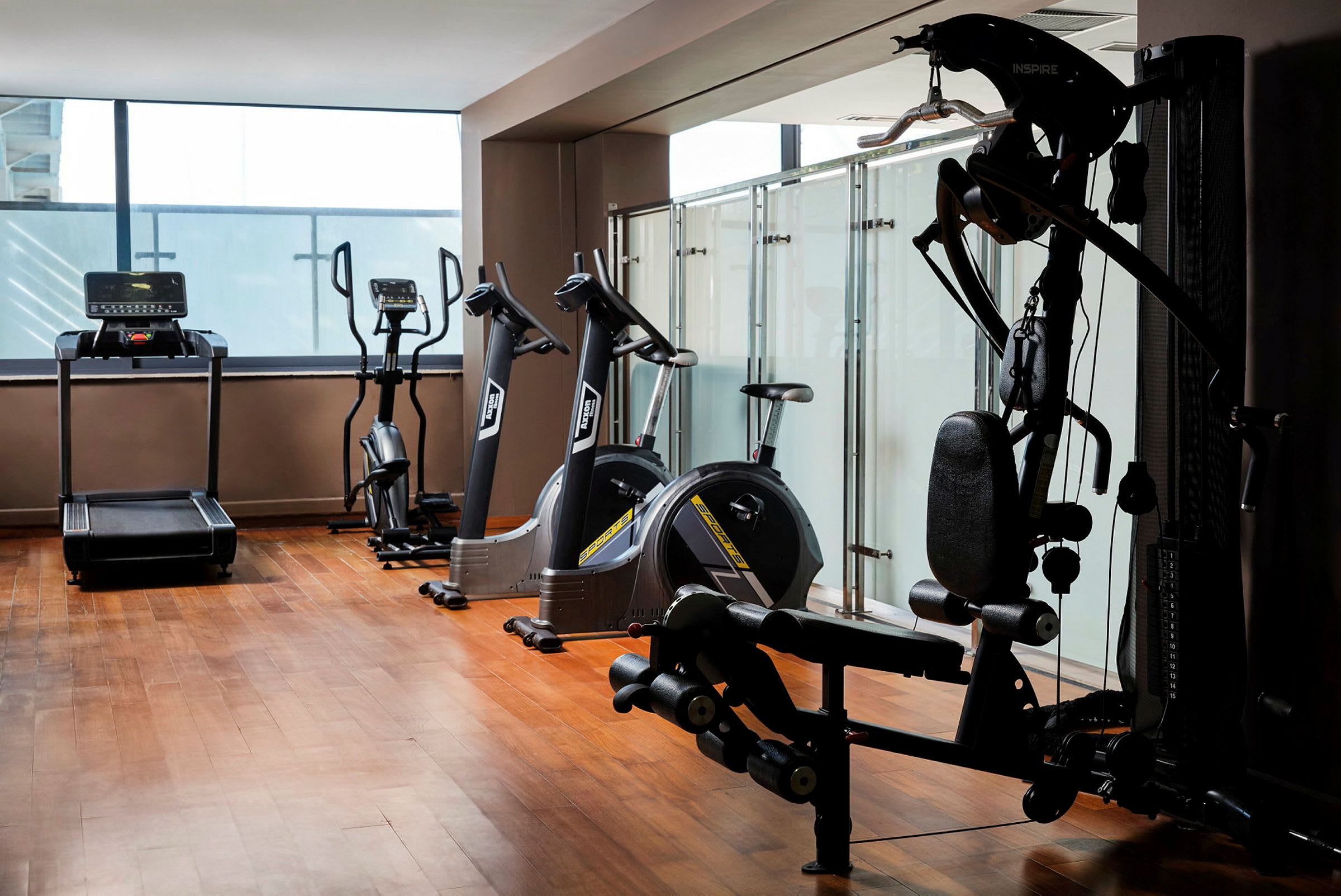 Ett gym med olika träningsmaskiner inklusive löpband, motionscyklar och styrketräningsutrustning, beläget i ett rum med trägolv och glasväggar. (AI-genererad text)