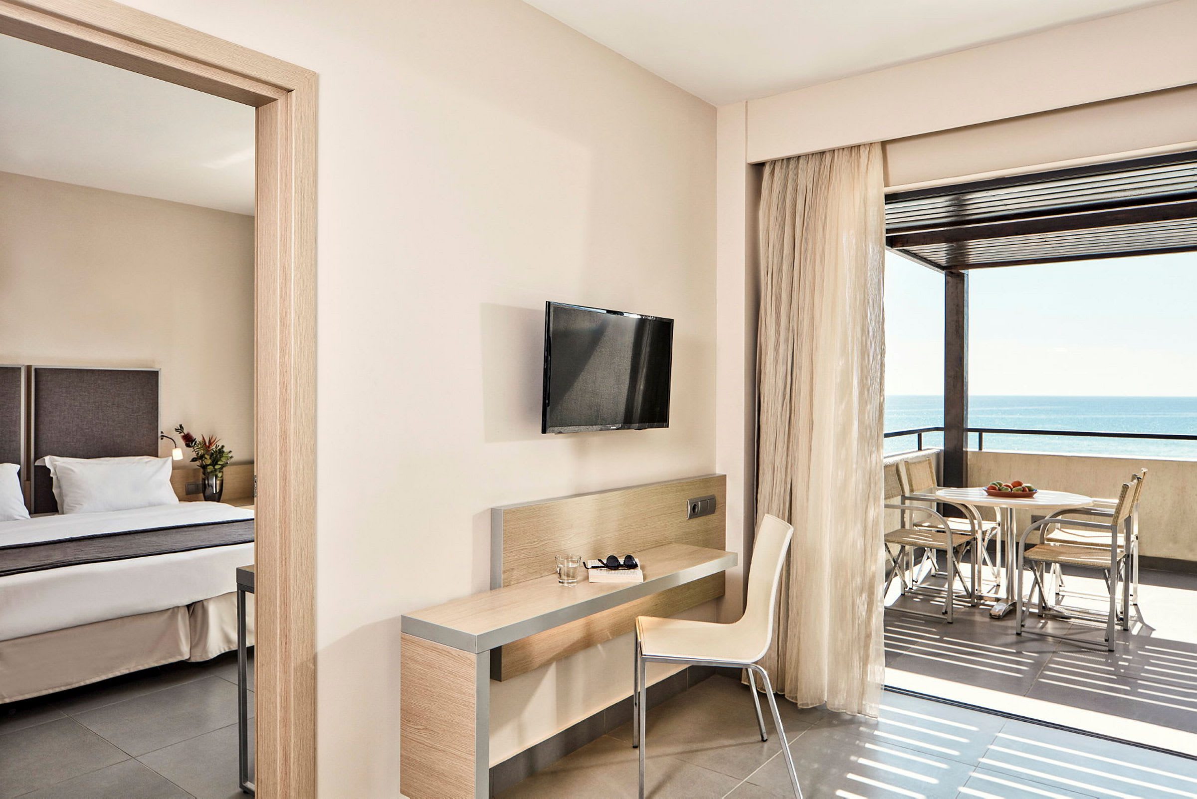 Ett hotellrum med en säng, skrivbord, stol, väggmonterad TV och balkong med utsikt över havet. (AI-genererad text)