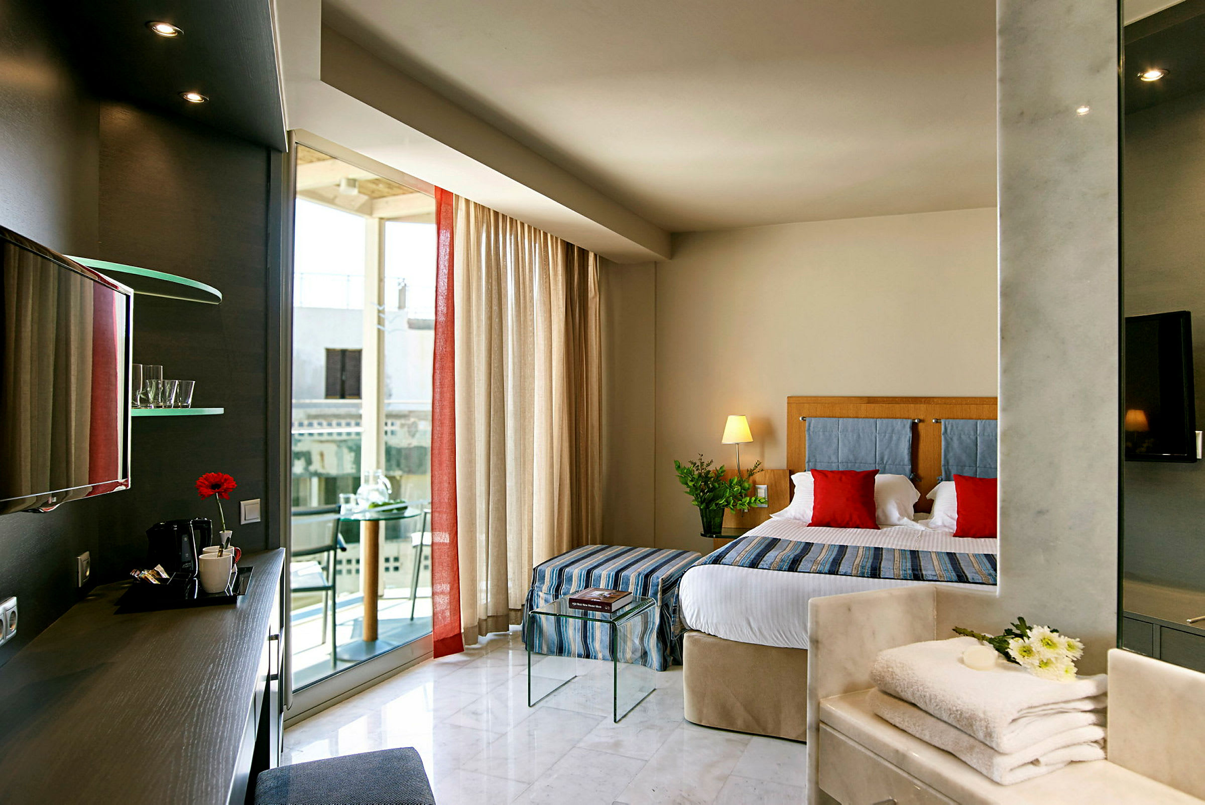 Ett hotellrum med en säng, dekorativa kuddar, ett skrivbord med kaffe och röd blomma, samt ett glasbord med böcker och balkong i bakgrunden. (AI-genererad text)