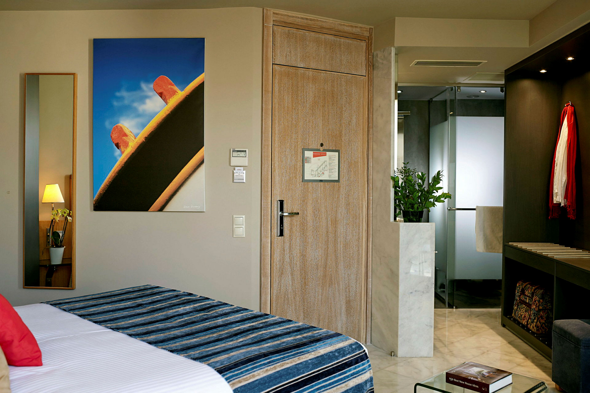 Ett hotellrum med en säng i förgrunden, en trädörr i mitten, och en spegel samt en tavla på väggen. I bakgrunden syns ett badrum med moderna detaljer. (AI-genererad text)