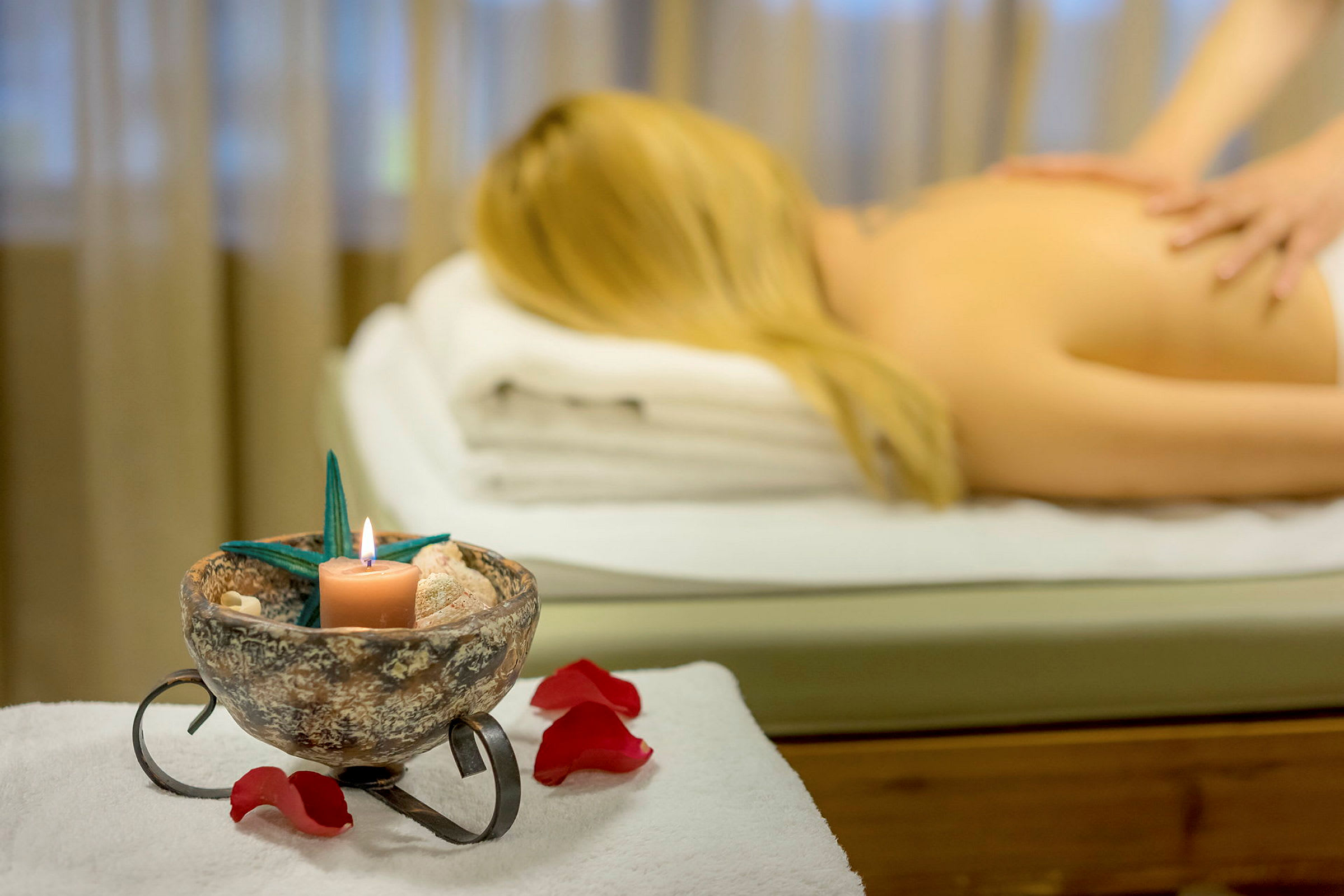 En massagebänk med en person liggandes under behandling. En dekoration med ett tänt ljus och blommor i förgrunden. (AI-genererad text)