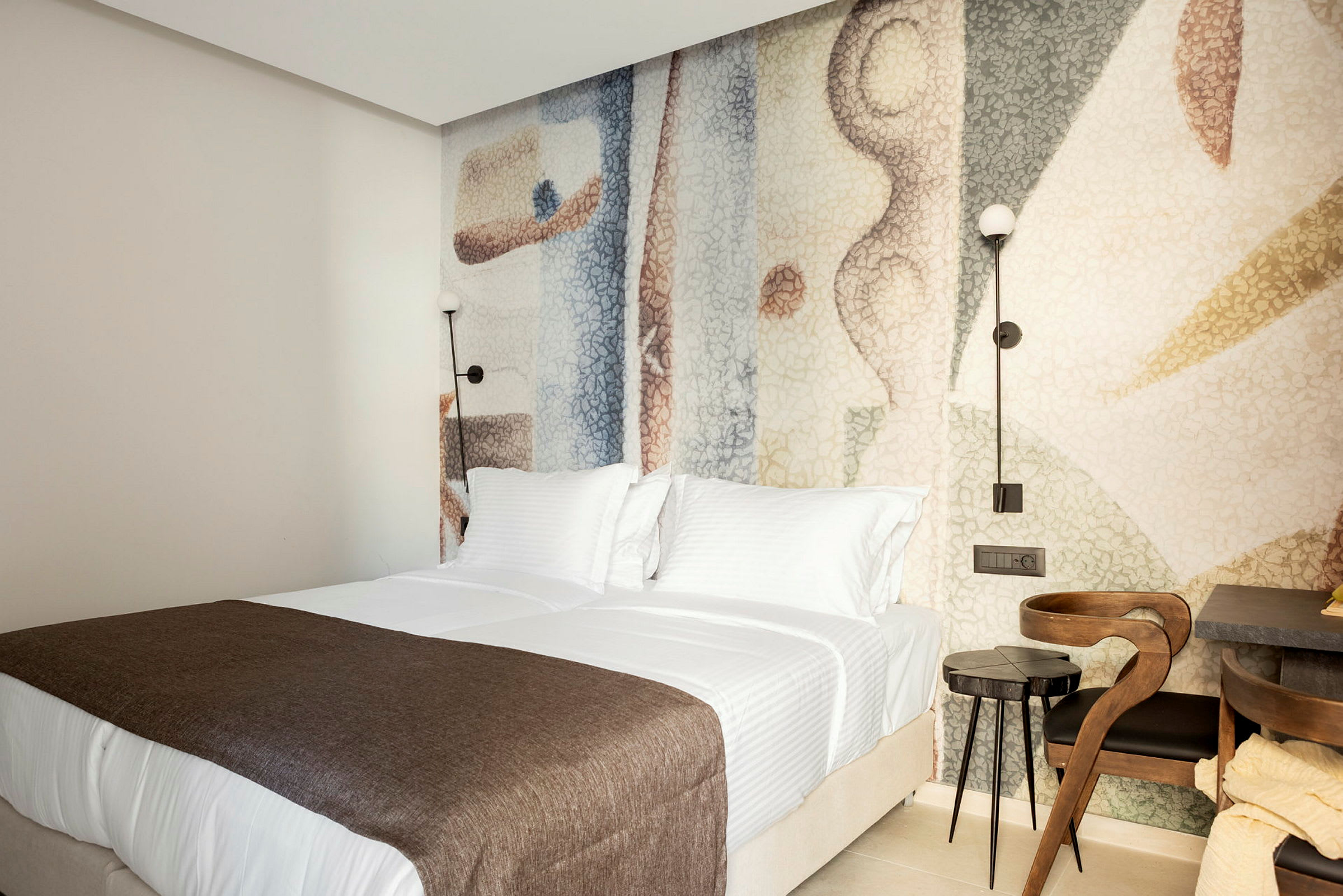 Ett hotellrum med två sängar täckta av vita sängkläder och bruna överkast. Sänggaveln har en dekorativ färgglad bakgrund. (AI-genererad text)