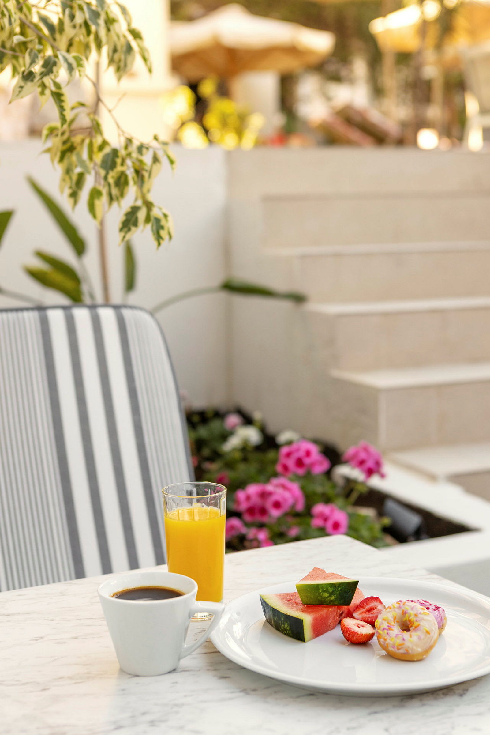 Ett bord med en tallrik som innehåller vattenmelonbitar, jordgubbar och en munk bredvid en kopp kaffe och ett glas apelsinjuice. I bakgrunden syns blommor och trappor. (AI-genererad text)
