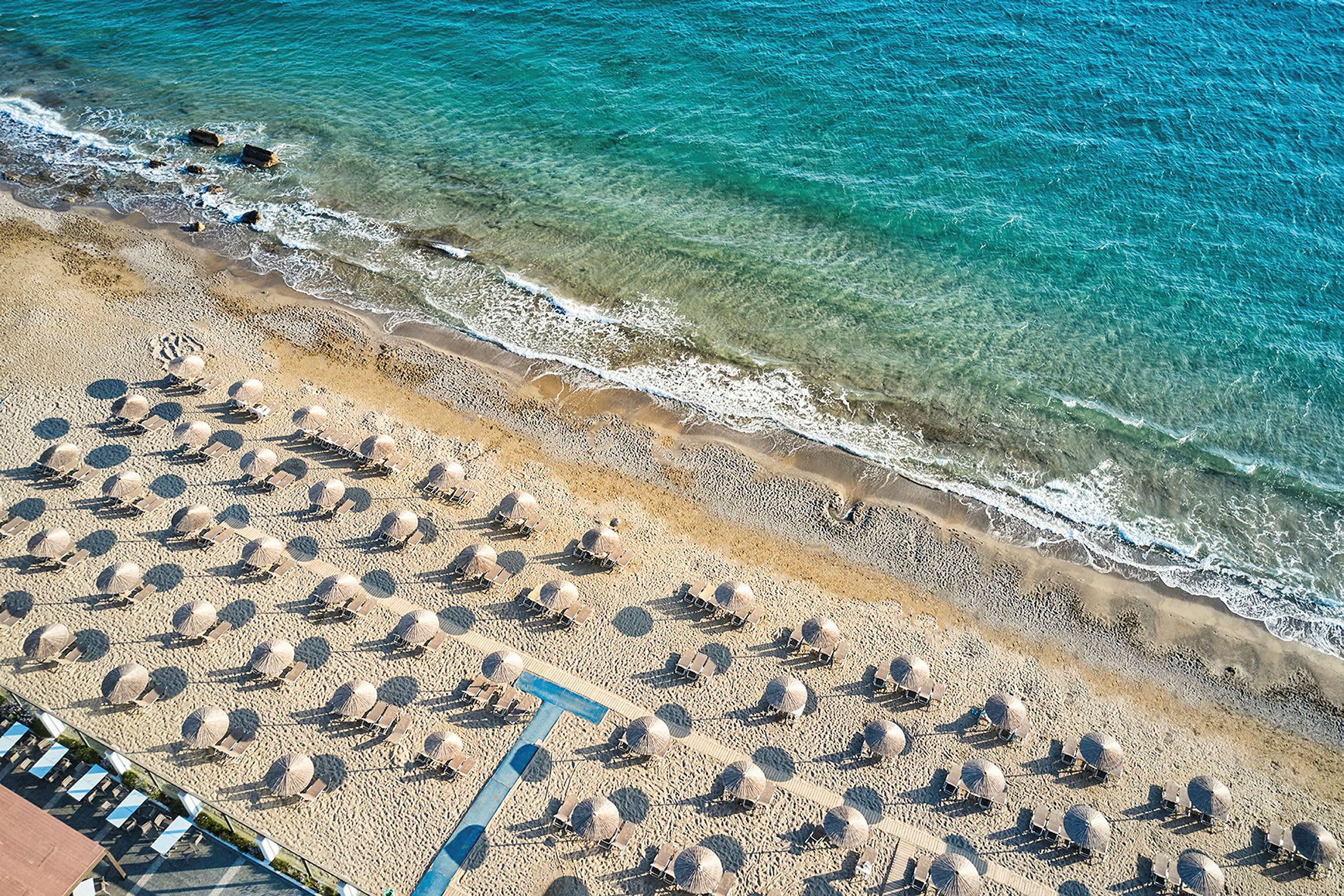 Flygfoto av en sandstrand med solstolar och parasoller, med kustlinjen och blått hav i bakgrunden. (AI-genererad text)