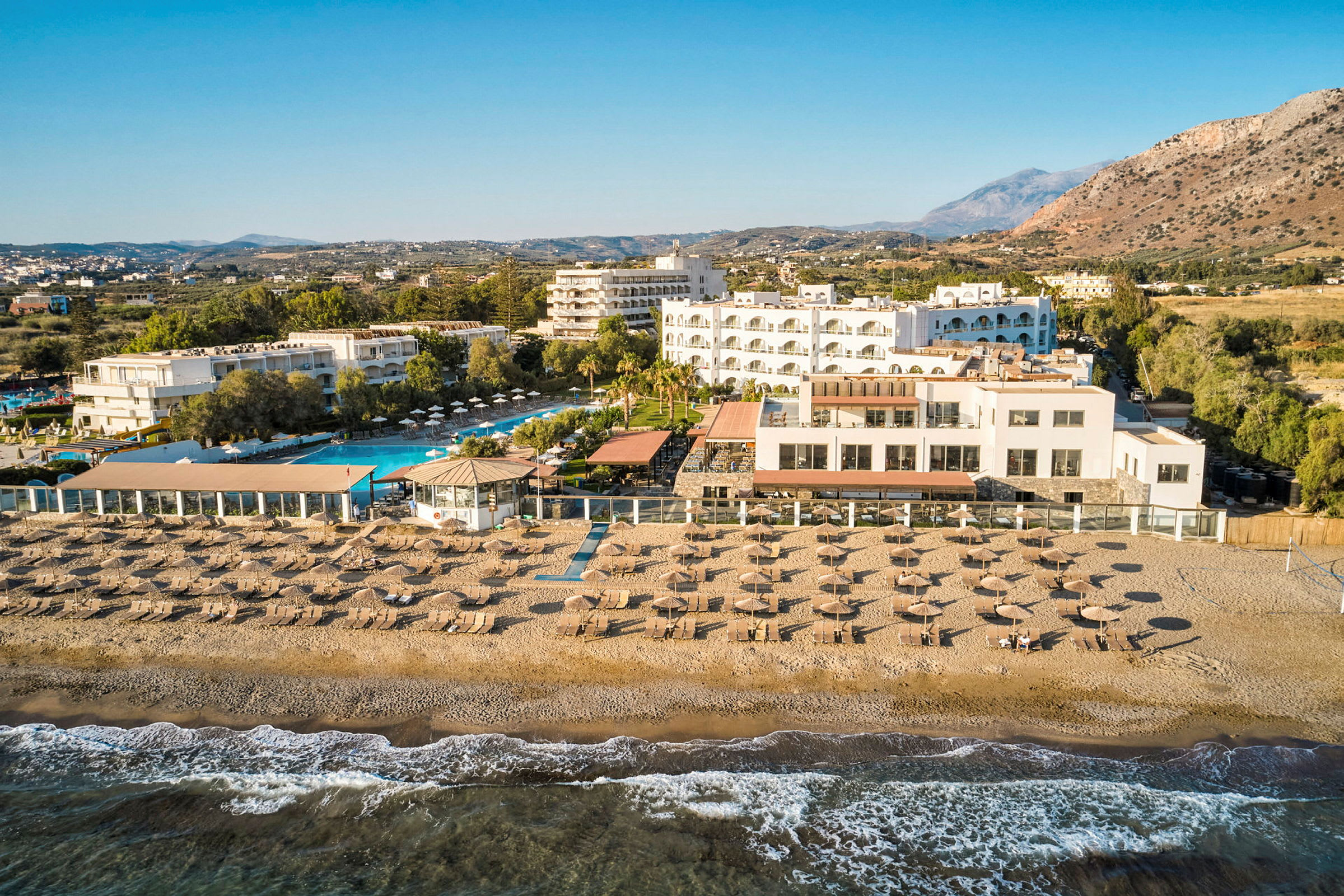 Ett hotellkomplex vid stranden med poolområde, solstolar, parasoller och en terrass med utsikt över havet. (AI-genererad text)