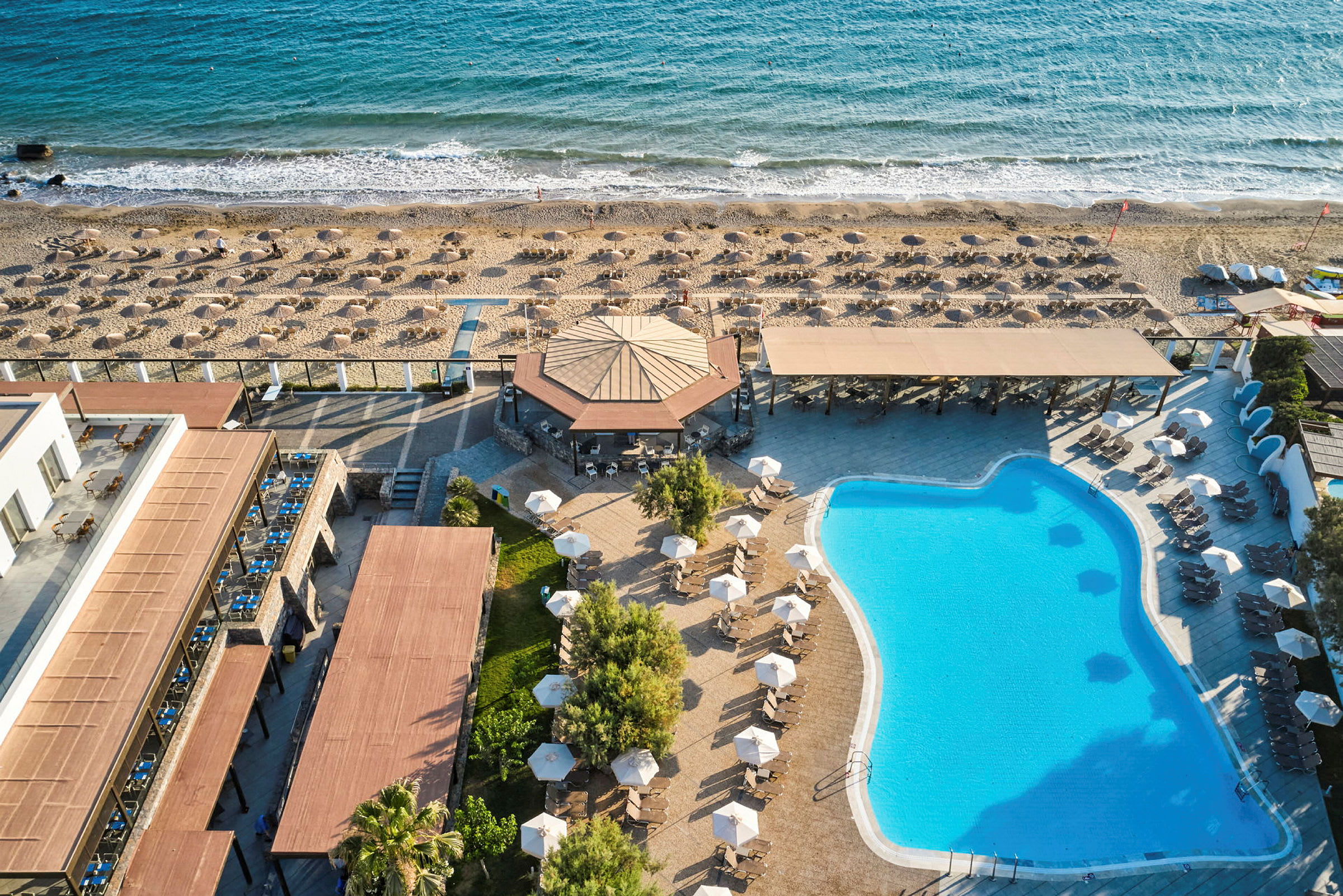 Flygfoto av en semesteranläggning med pool, solstolar och parasoll nära en strand. (AI-genererad text)