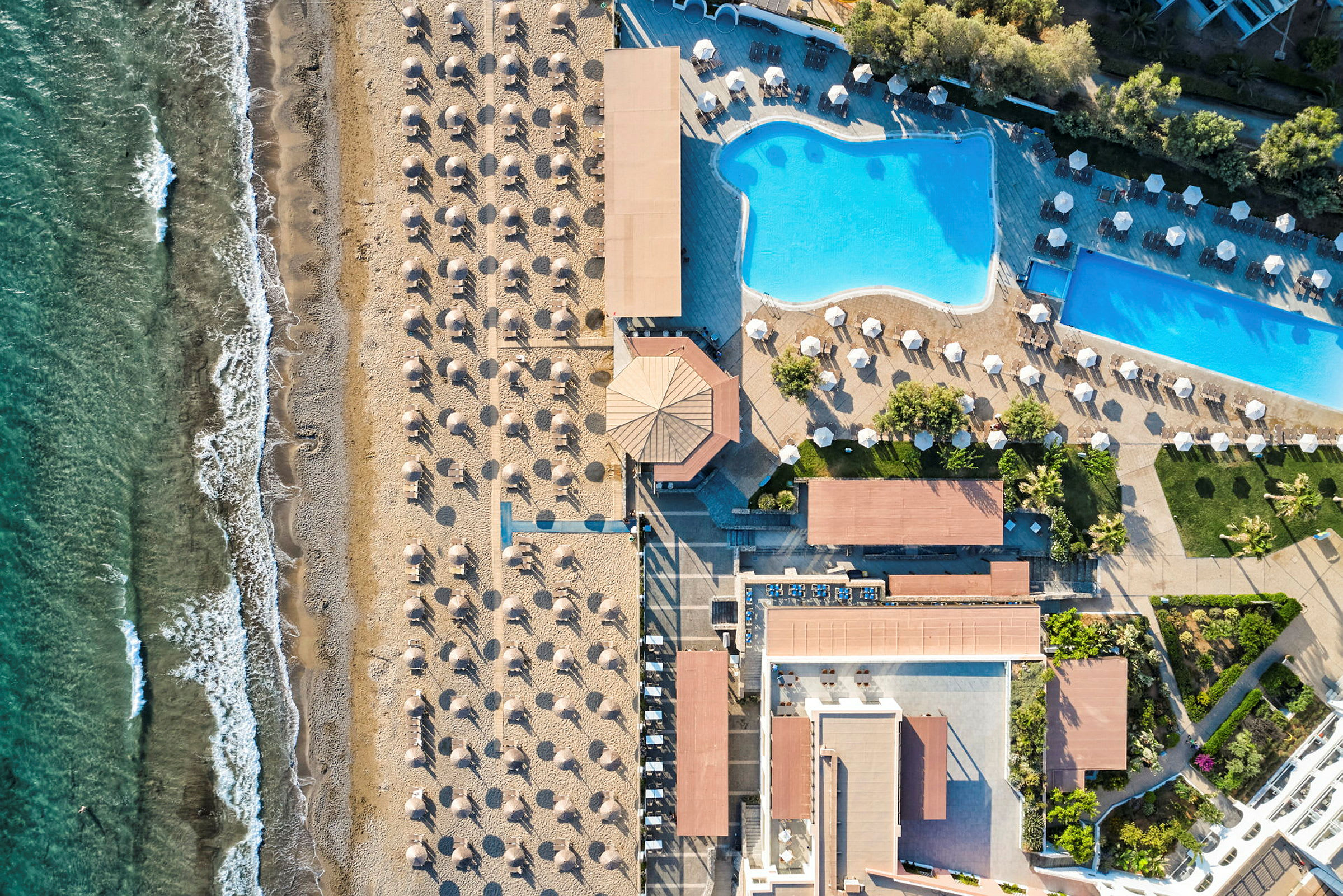 Flygfotografi av en strand med solstolar och parasoller bredvid en stor pool och semesteranläggning. (AI-genererad text)