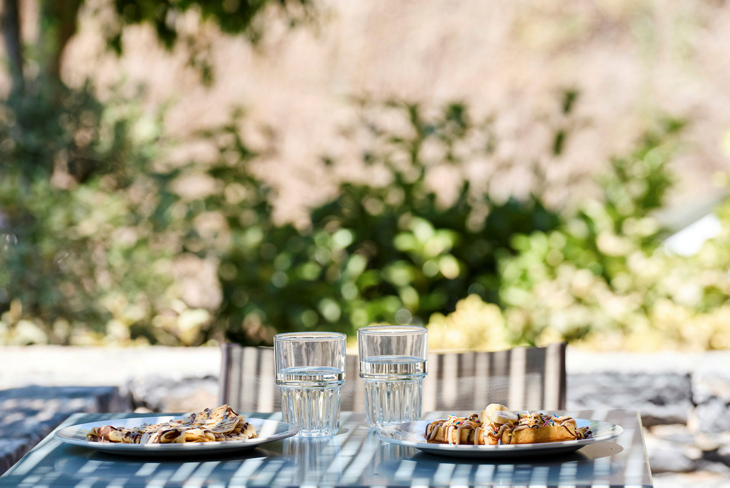 Två tallrikar med mat och två glas vatten står på ett blått bord utomhus, med grönska i bakgrunden. (AI-genererad text)