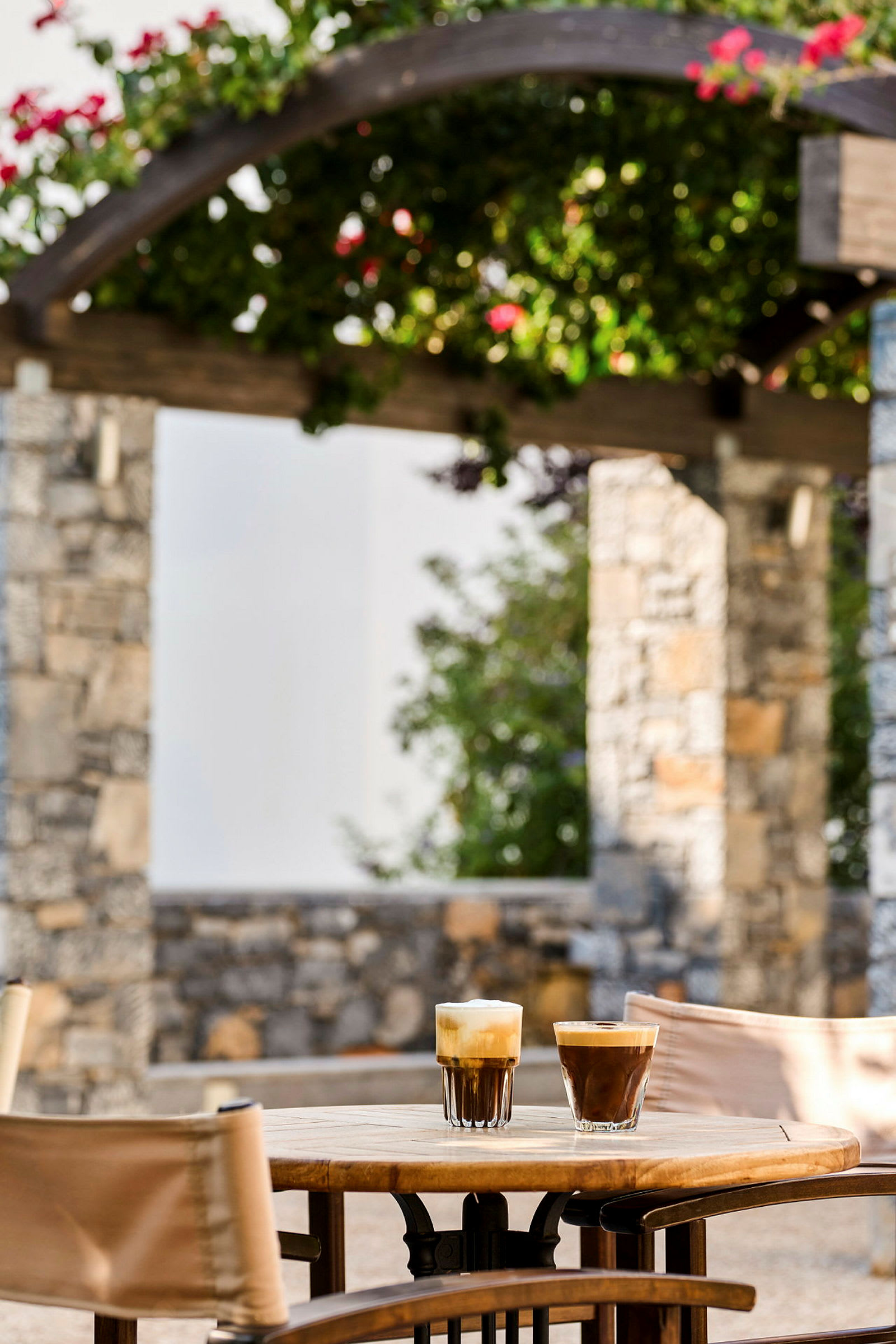 Två glas med kaffe står på ett bord utomhus, med en stenstruktur och blommande växter i bakgrunden. (AI-genererad text)