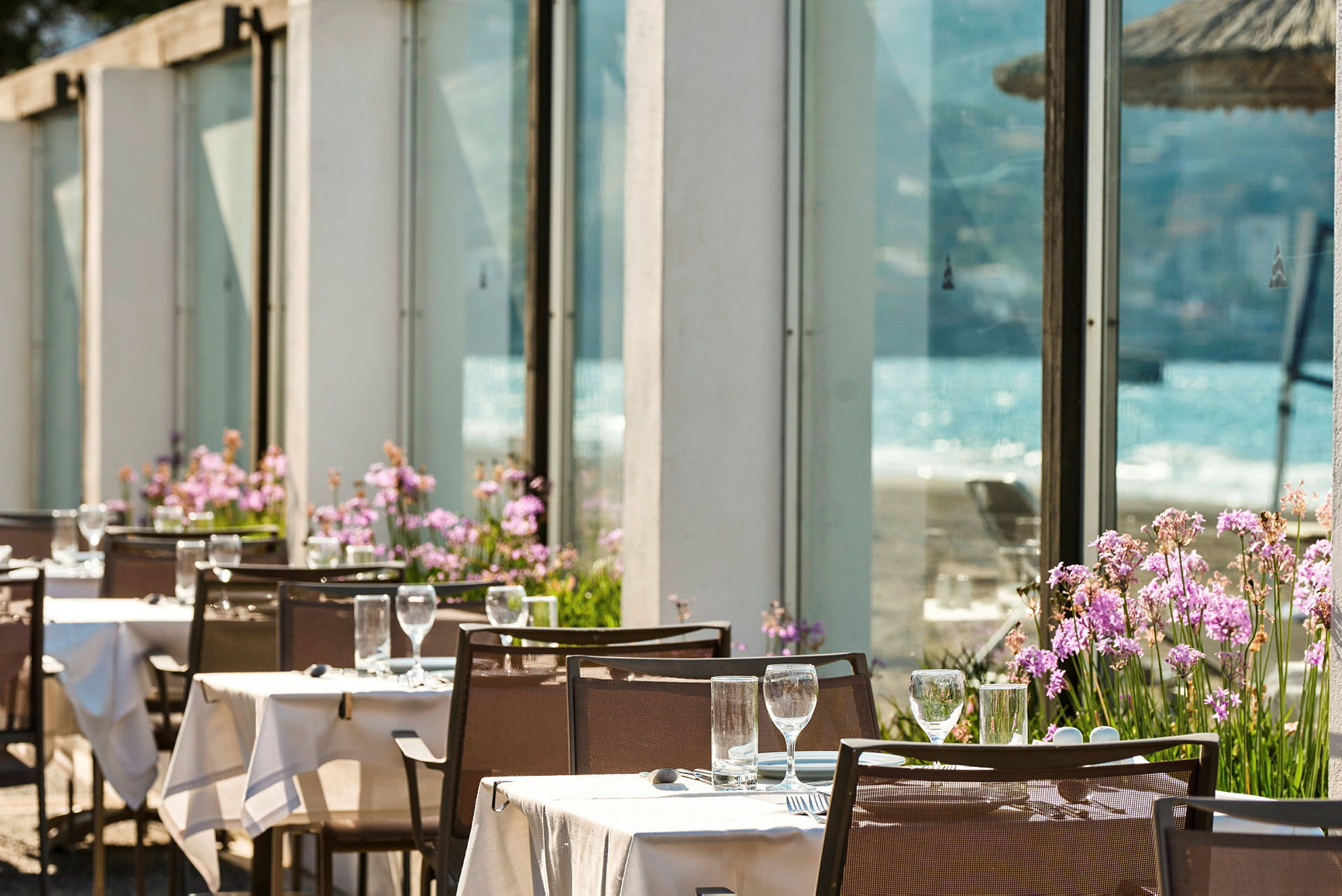 Uteservering med dukade bord, utsmyckade med glas och bestick, omgivna av färgglada blommor nära fönsterrutor. I bakgrunden syns havet. (AI-genererad text)