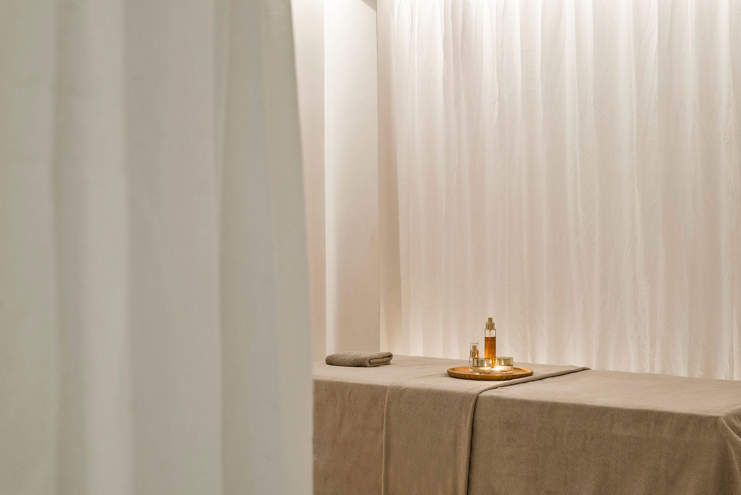 Ett spa-rum med en massagebänk och ljusa gardiner. På bänken finns en bricka med oljor och små glas. (AI-genererad text)