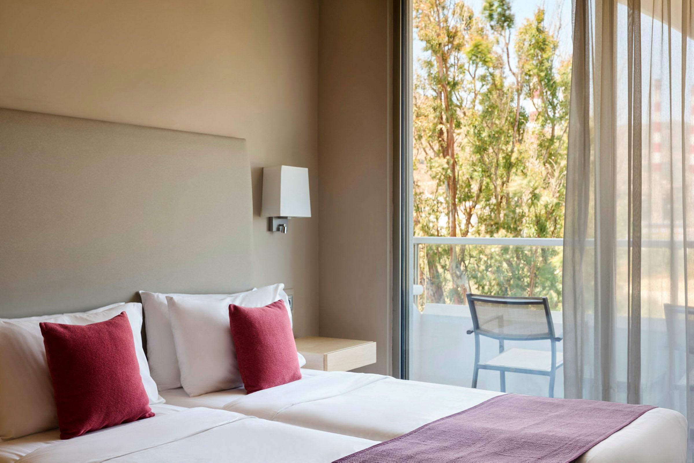 Hotellrum med två sängar, röda kuddar och utsikt mot en balkong med stolar och träd utanför. (AI-genererad text)