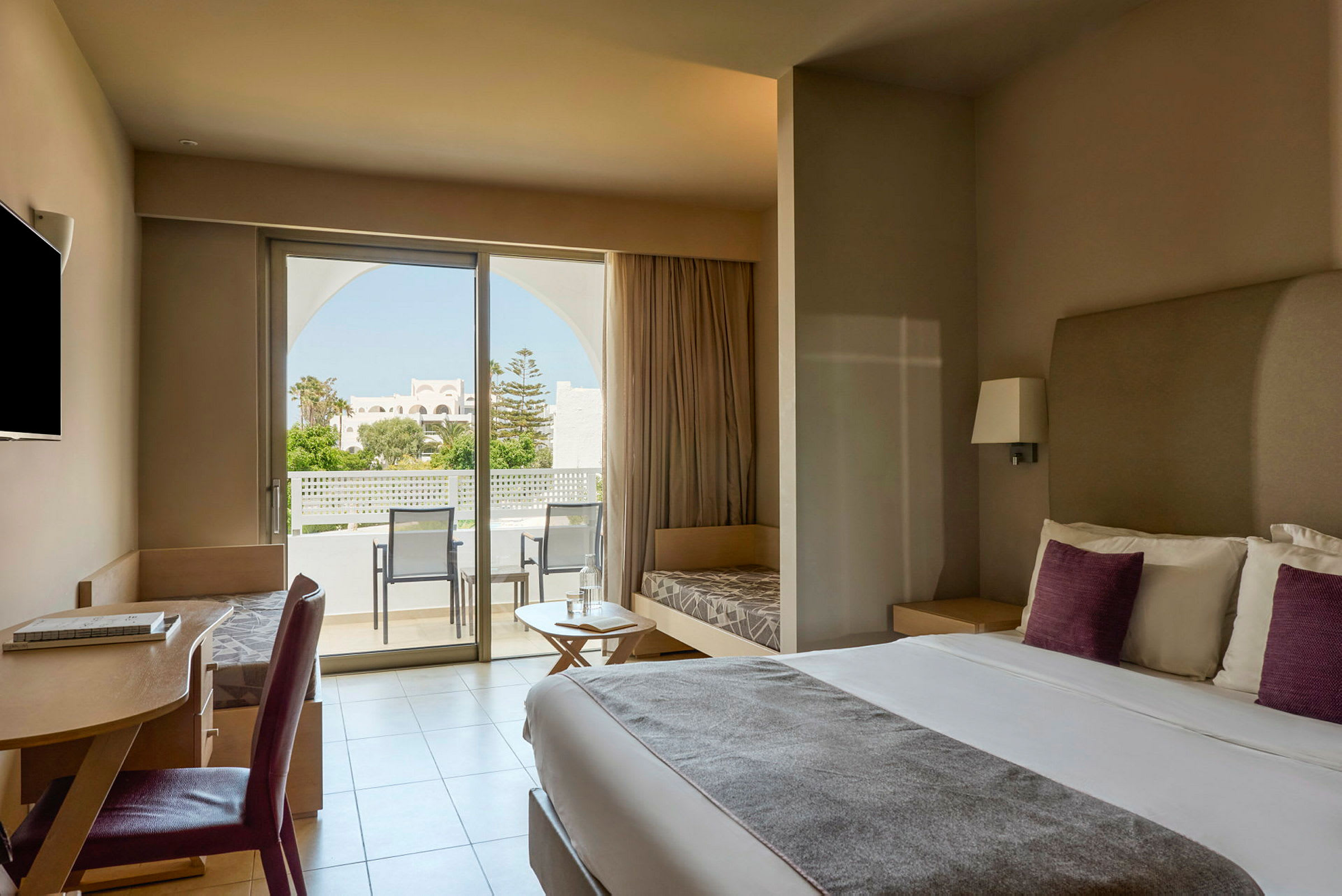 Ett modernt hotellrum med en dubbelsäng, skrivbord, sittmöbler och ett stort fönster som leder ut till en balkong med utsikt över en trädgård. (AI-genererad text)
