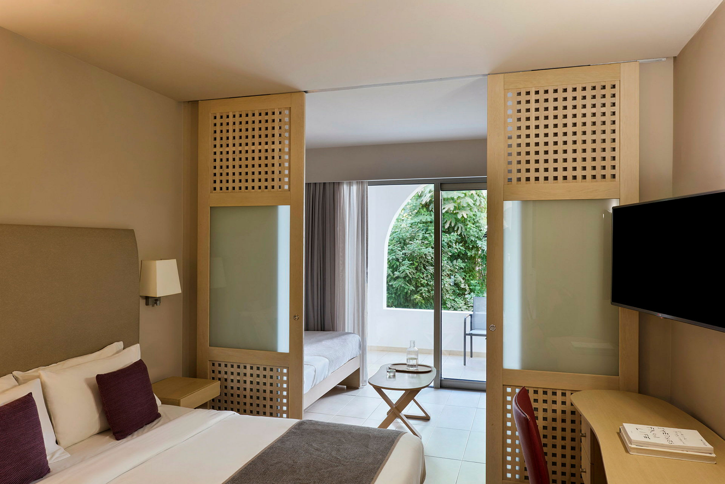 Hotellrum med dubbelsäng, sittmöbler och glasdörr som leder till en balkong med utsikt över grönska. (AI-genererad text)