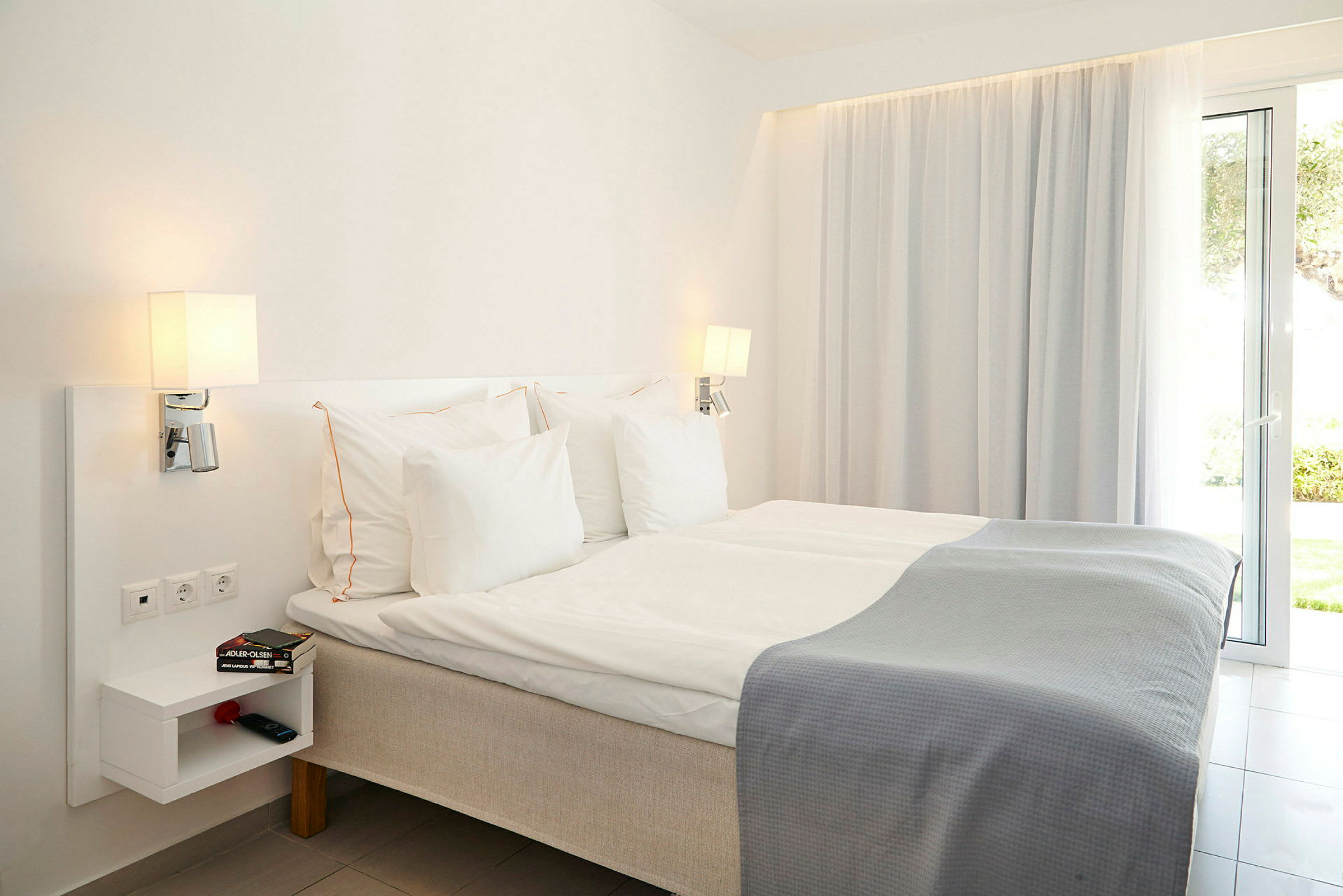 Ett hotellrum med en säng, vita sängkläder och grå filt med lampor, hylla och fönster. (AI-genererad text)