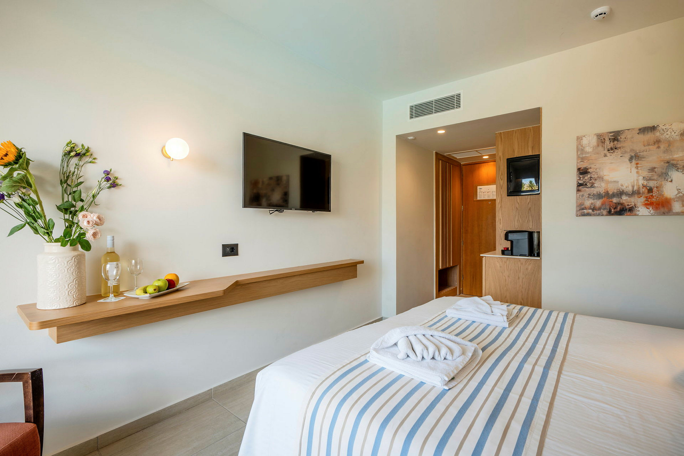 Hotellrum med en bäddad säng, vägg-TV, och hylla med dekorerande blommor och frukt. (AI-genererad text)