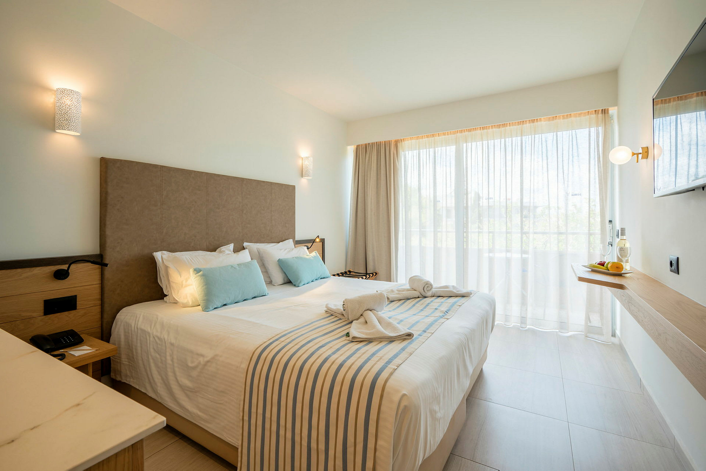 Ett hotellrum med en dubbelsäng, prydnad med kuddar och hoprullade handdukar, fönster med gardiner samt en väggmonterad hylla med en flaska och frukt. (AI-genererad text)