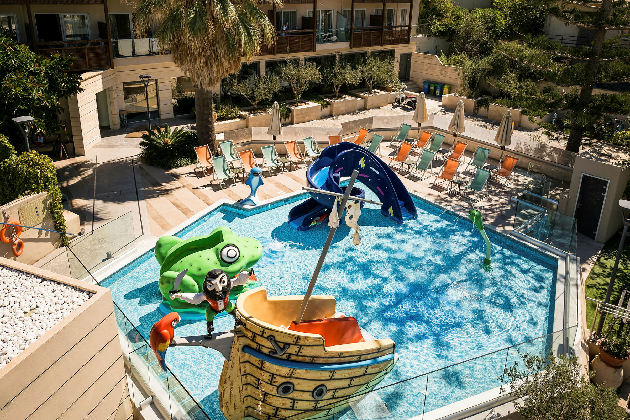 En pool med barnvänliga figurer och en rutschkana, omgiven av solstolar och en palm. (AI-genererad text)