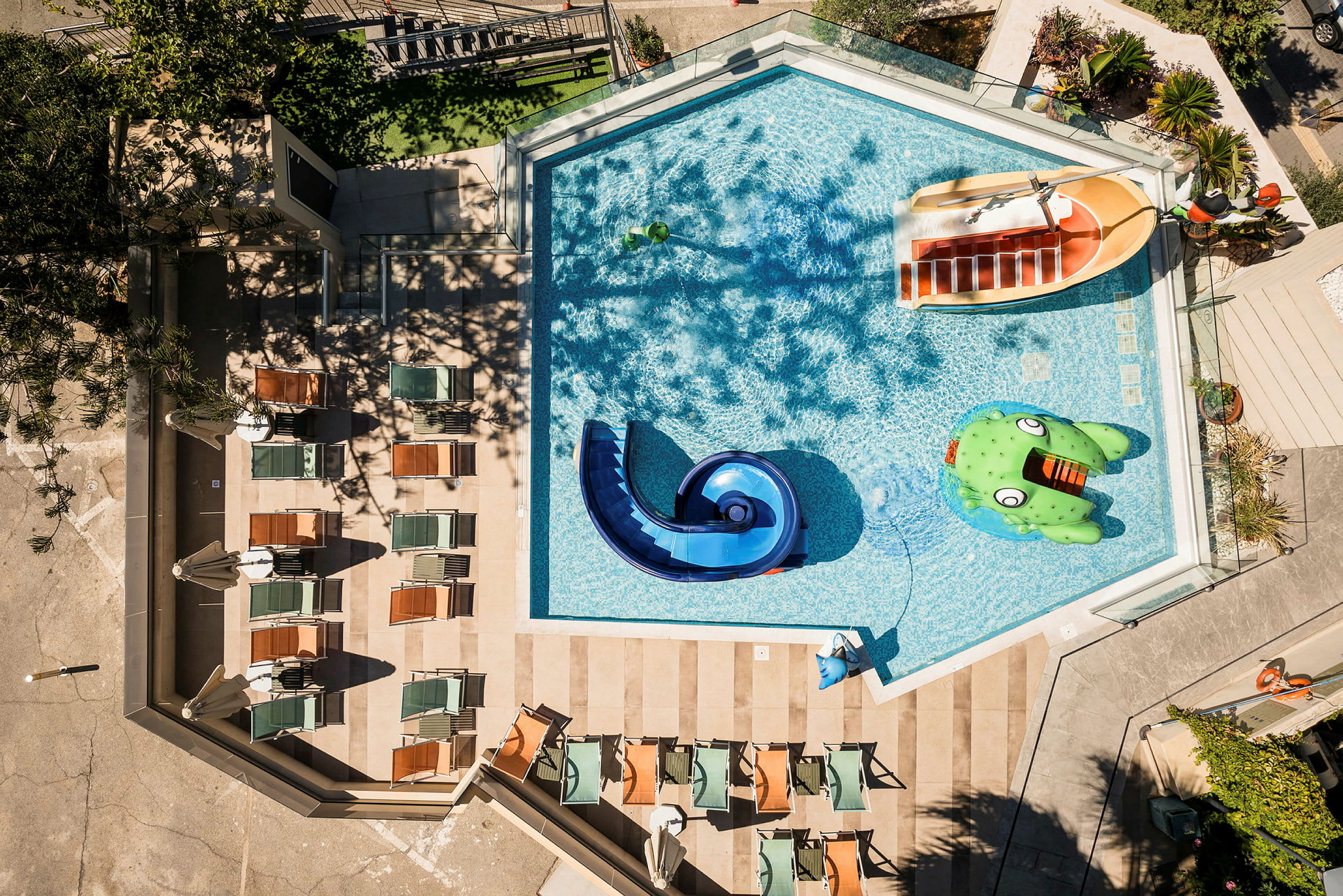 Fågelperspektiv över en pool med vattenrutschbana och uppblåsbara leksaker samt omgivande solstolar. (AI-genererad text)