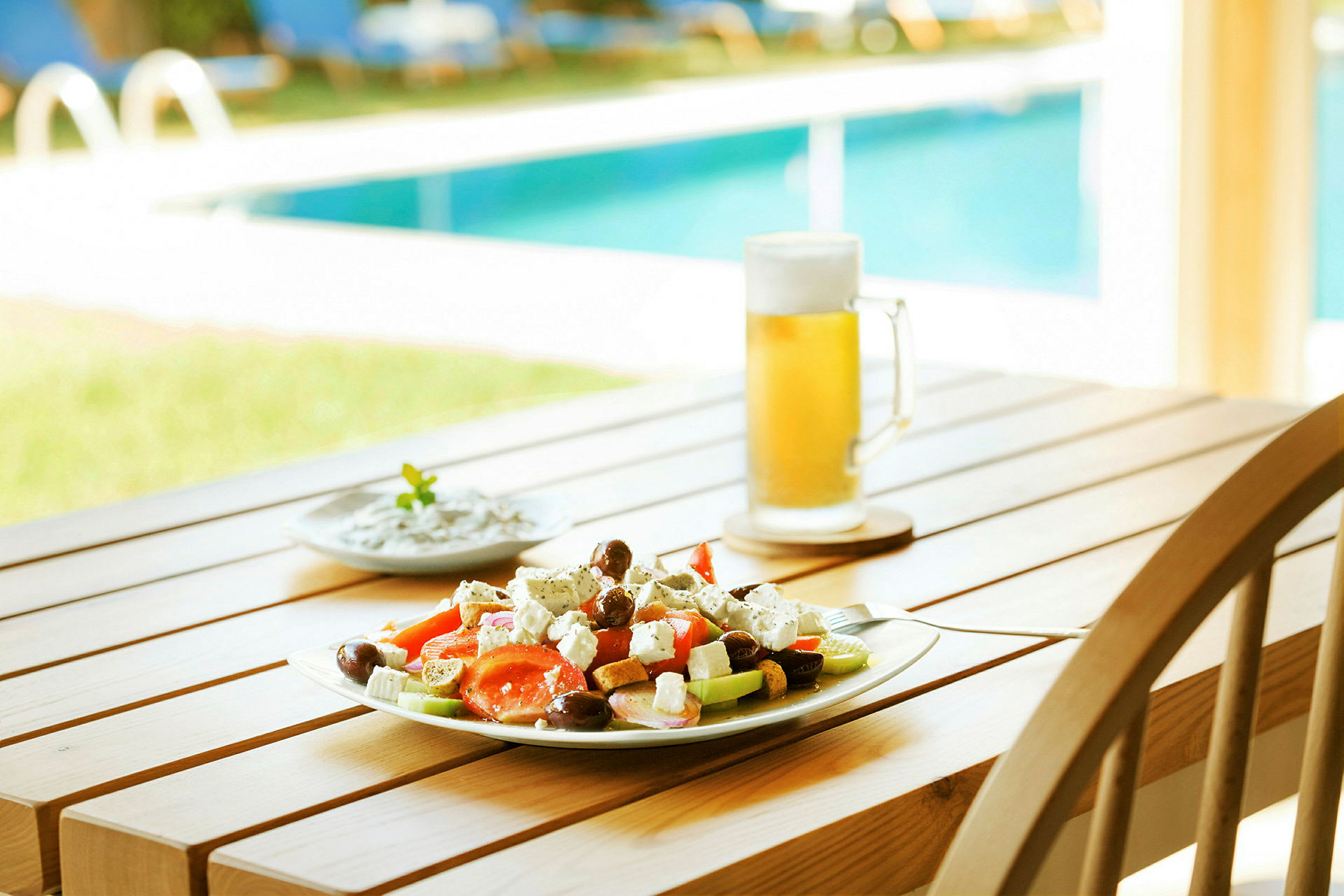 En tallrik med sallad, ett glas öl och en mindre skål står på ett bord nära en pool. (AI-genererad text)